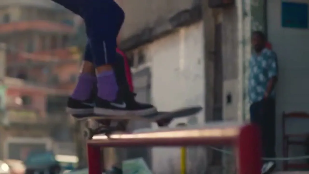 A young female skateboarder in a purple hoodie and dark pants grinds on a red rail in a vibrant, urban setting, with other people watching in the background.