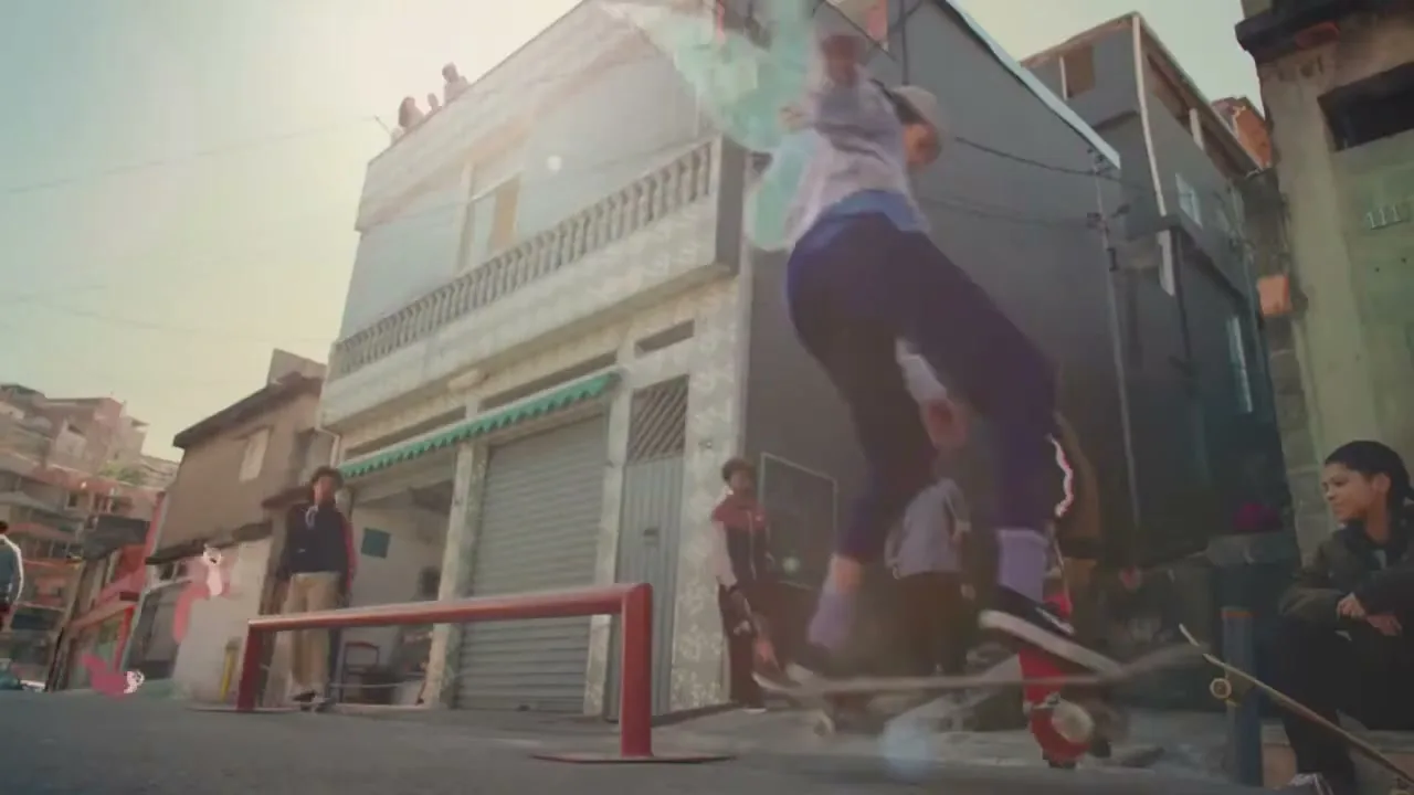 A young person with fairy wings skates on a rail, while other young people watch in a street with buildings in the background.