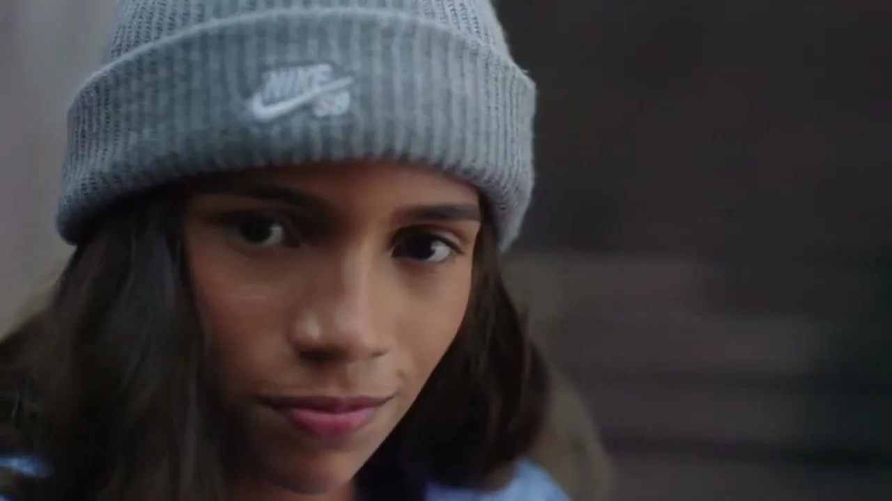 A young woman with long dark hair and a grey beanie with a Nike logo looks down, then up at the camera, then quickly away.