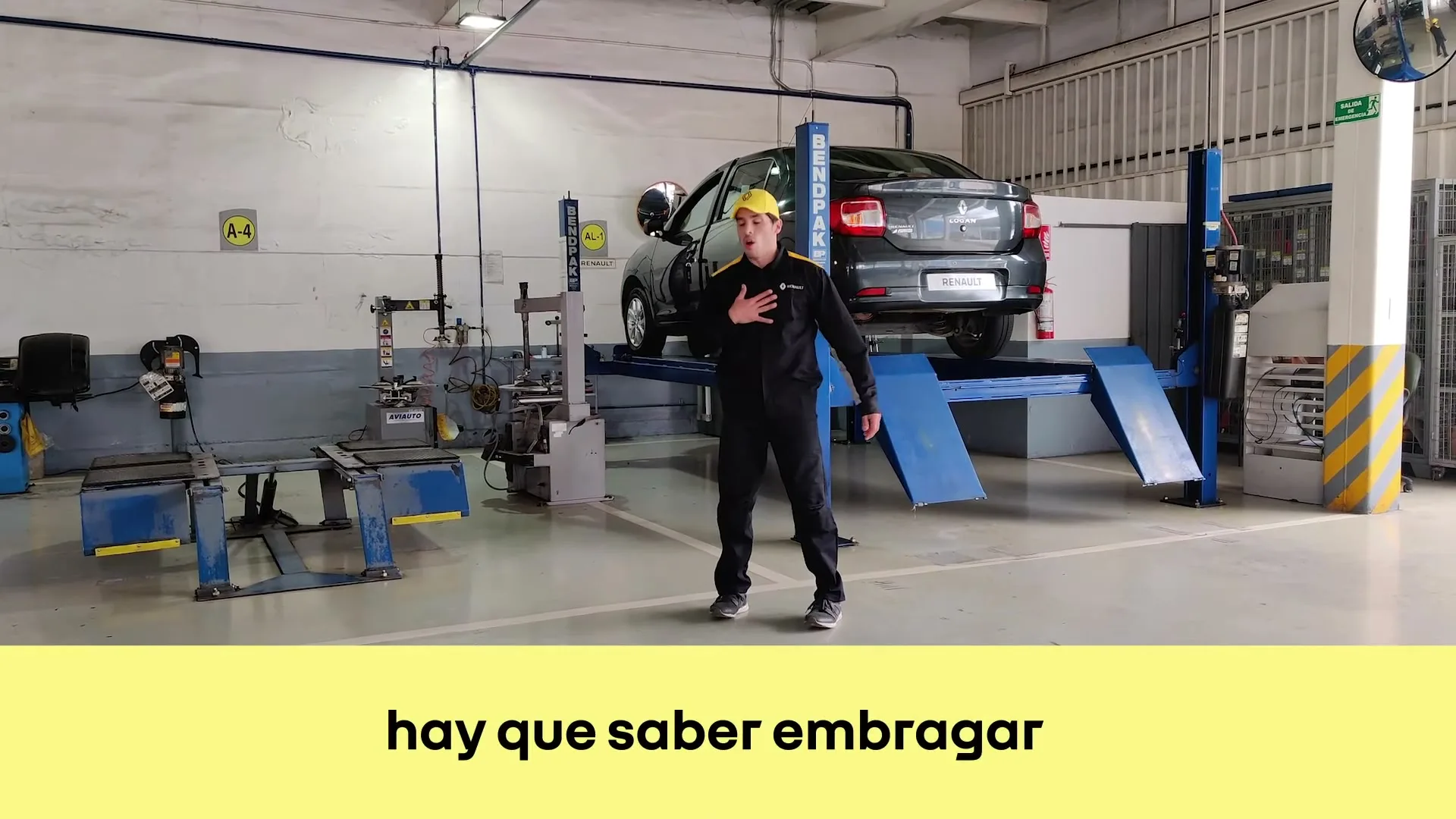 A male mechanic in a yellow cap and black uniform dances energetically in a car repair shop, with a dark grey car elevated on a blue lift behind him.