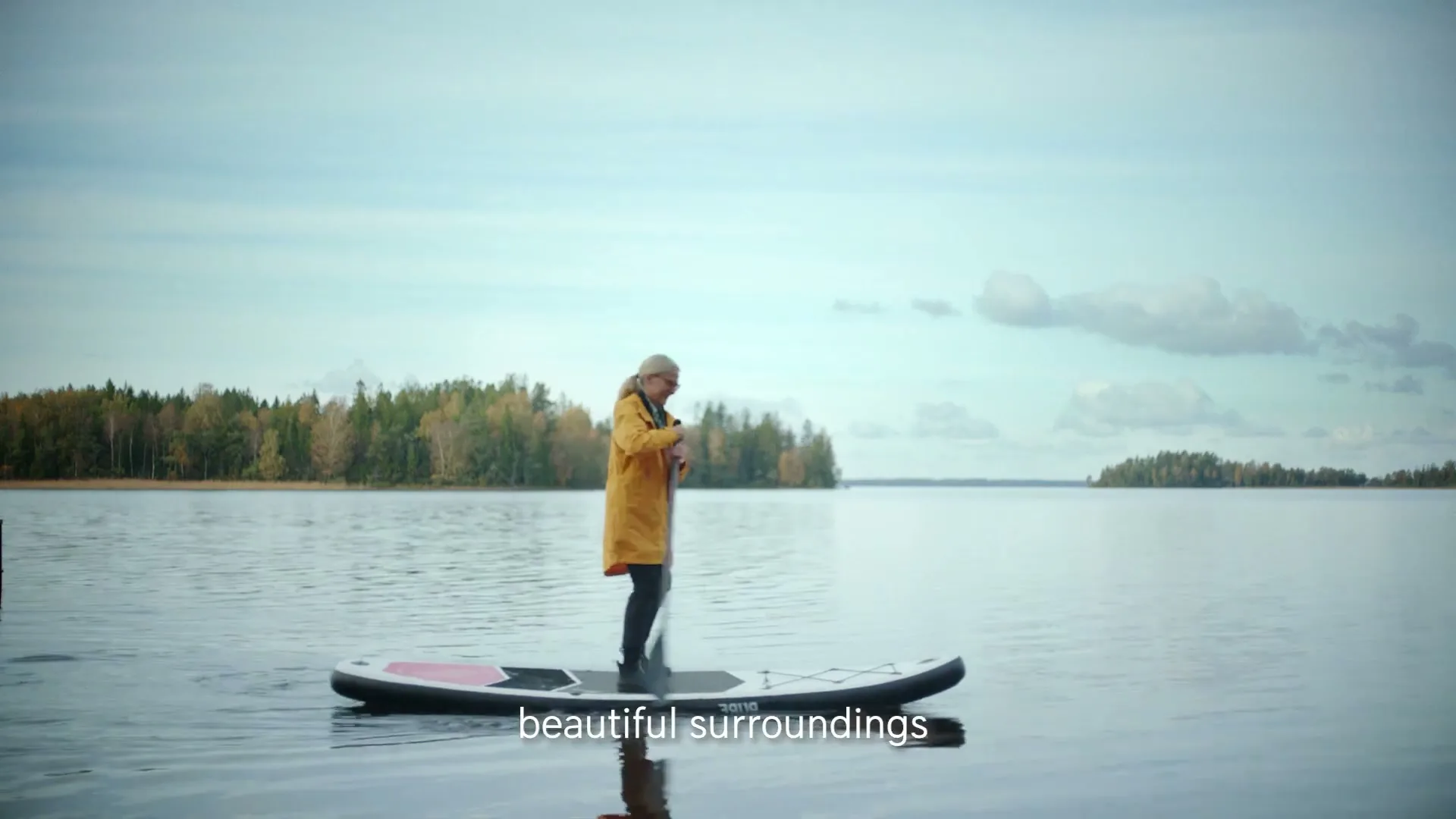 A woman in her 60s with blonde hair and a yellow raincoat stands on a paddleboard in a lake, paddling gently.