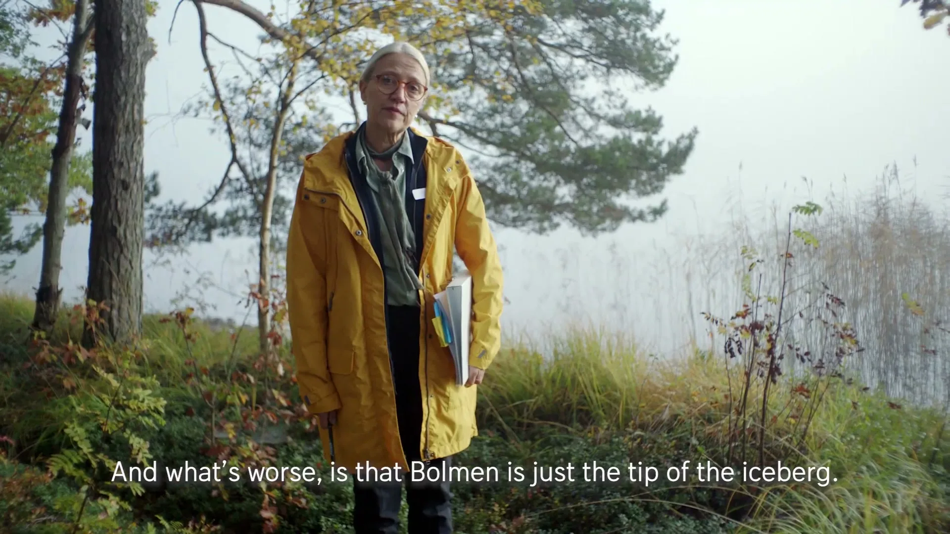 A blonde woman in her 50s, wearing a yellow raincoat and glasses, stands by a lake, holding a drawing and talking to the camera.