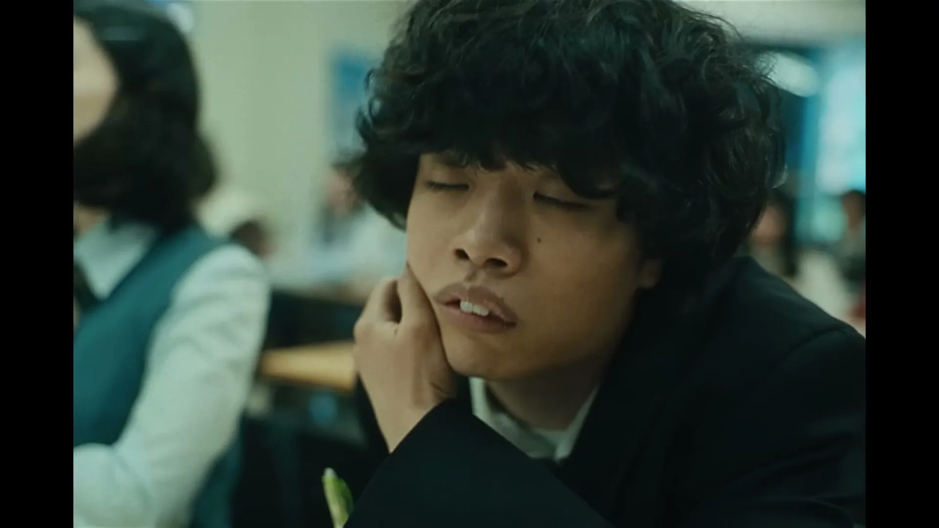 A young male student with curly hair, wearing a black blazer, rests his head on his hand, looking tired in a classroom setting.