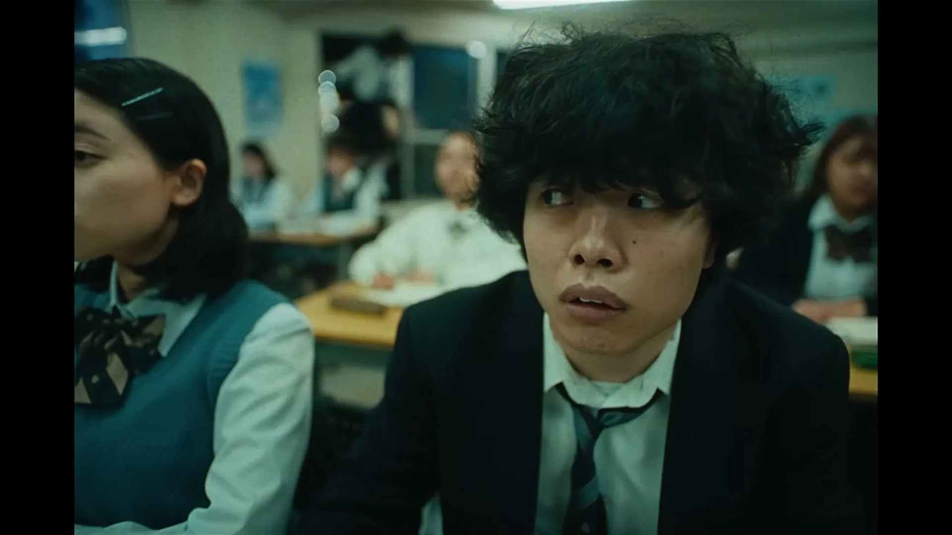 A male student with messy hair looks around nervously in a classroom, while a female student sits beside him, looking down.