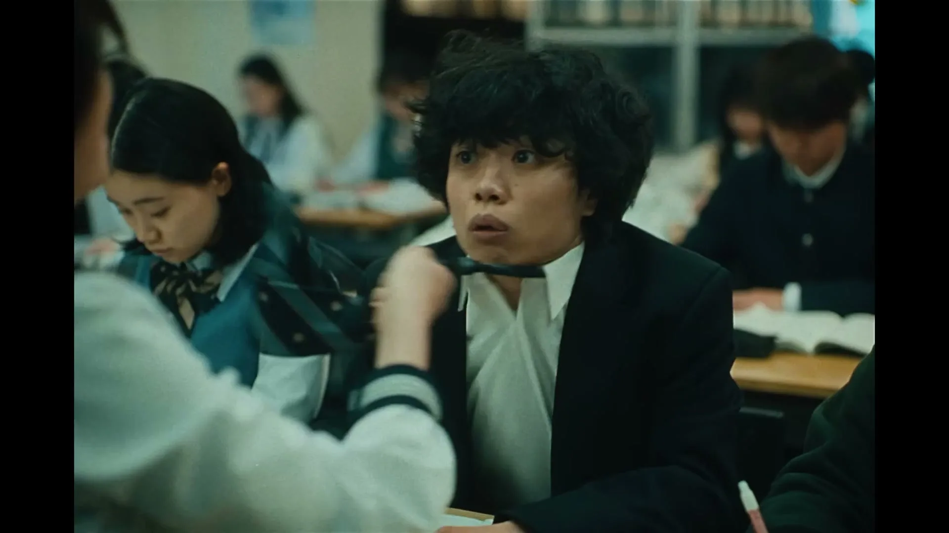 A female student in a white uniform pulls the tie of a male student with curly hair, who looks surprised in a classroom.