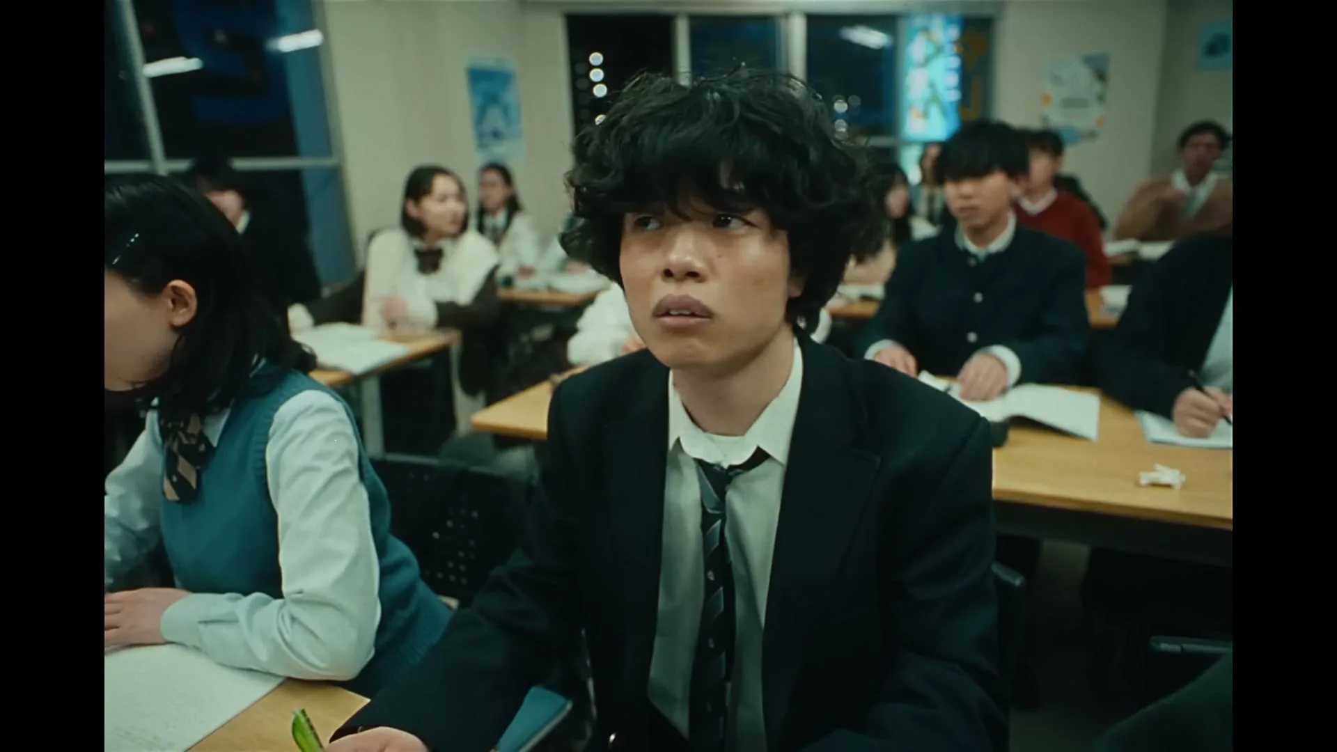 A male student with curly hair in a school uniform looks forward intently while sitting at a desk in a classroom full of other students.