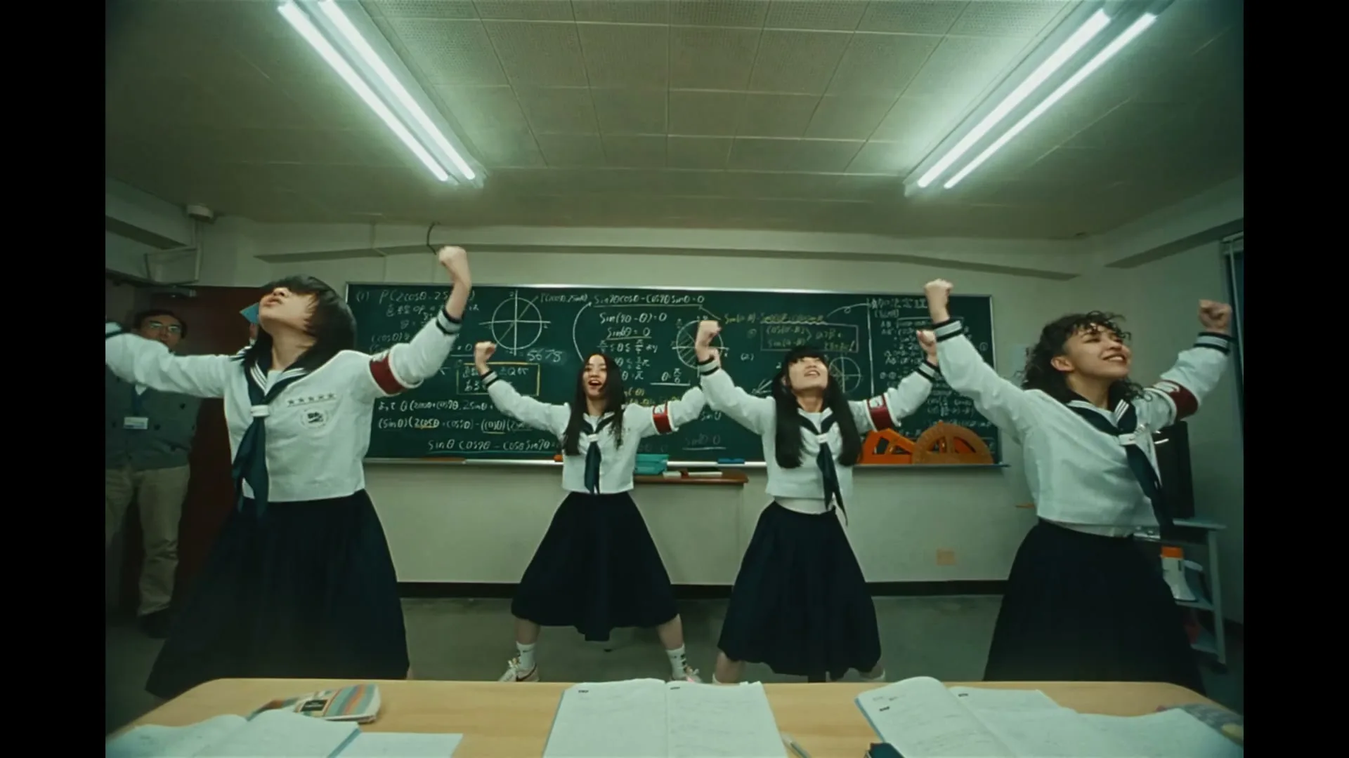 Four female students in sailor uniforms dance energetically in a classroom, while a male teacher watches them from the background.