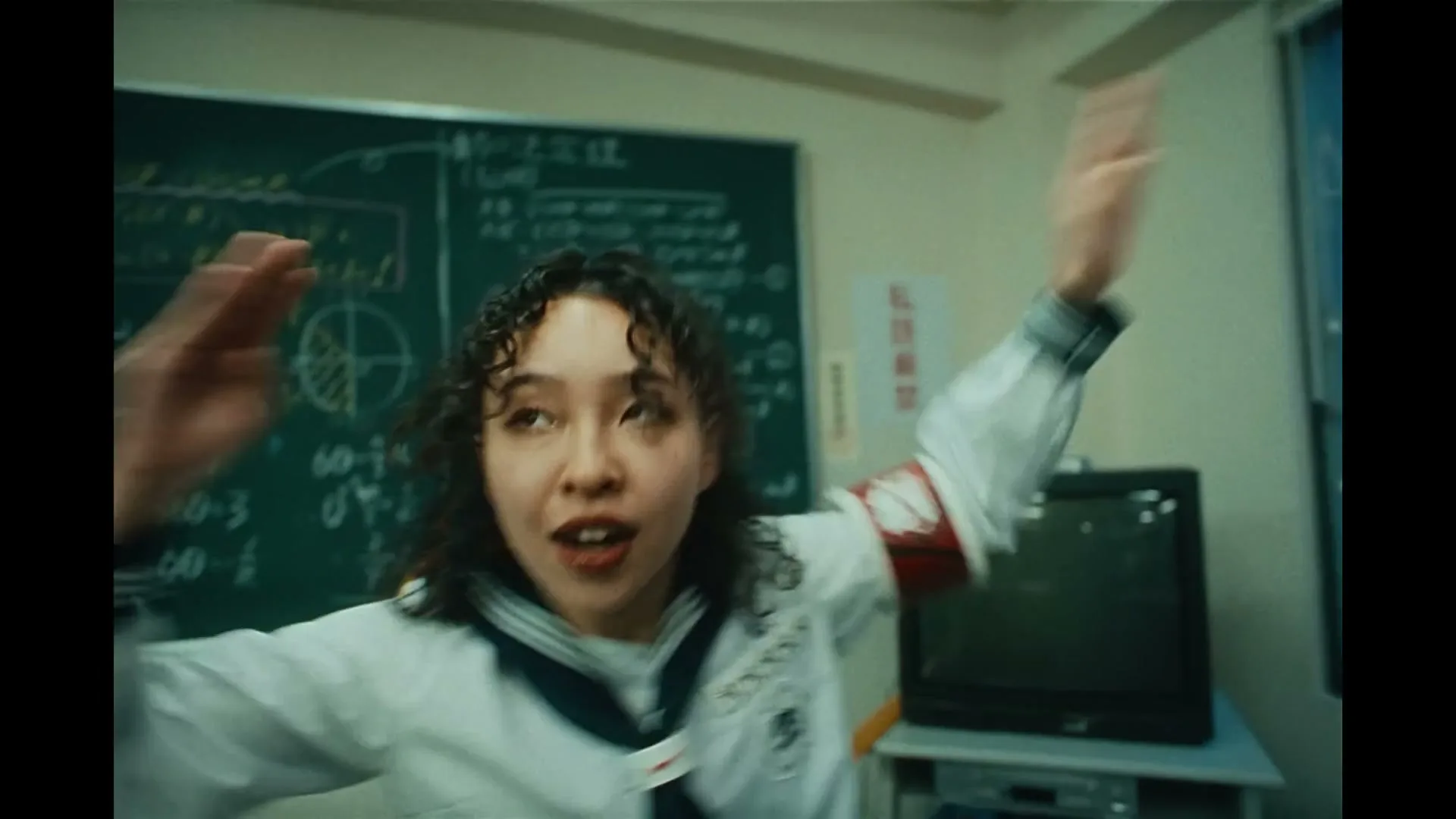 A young female student in a sailor uniform dances energetically in a classroom with a blackboard full of equations.