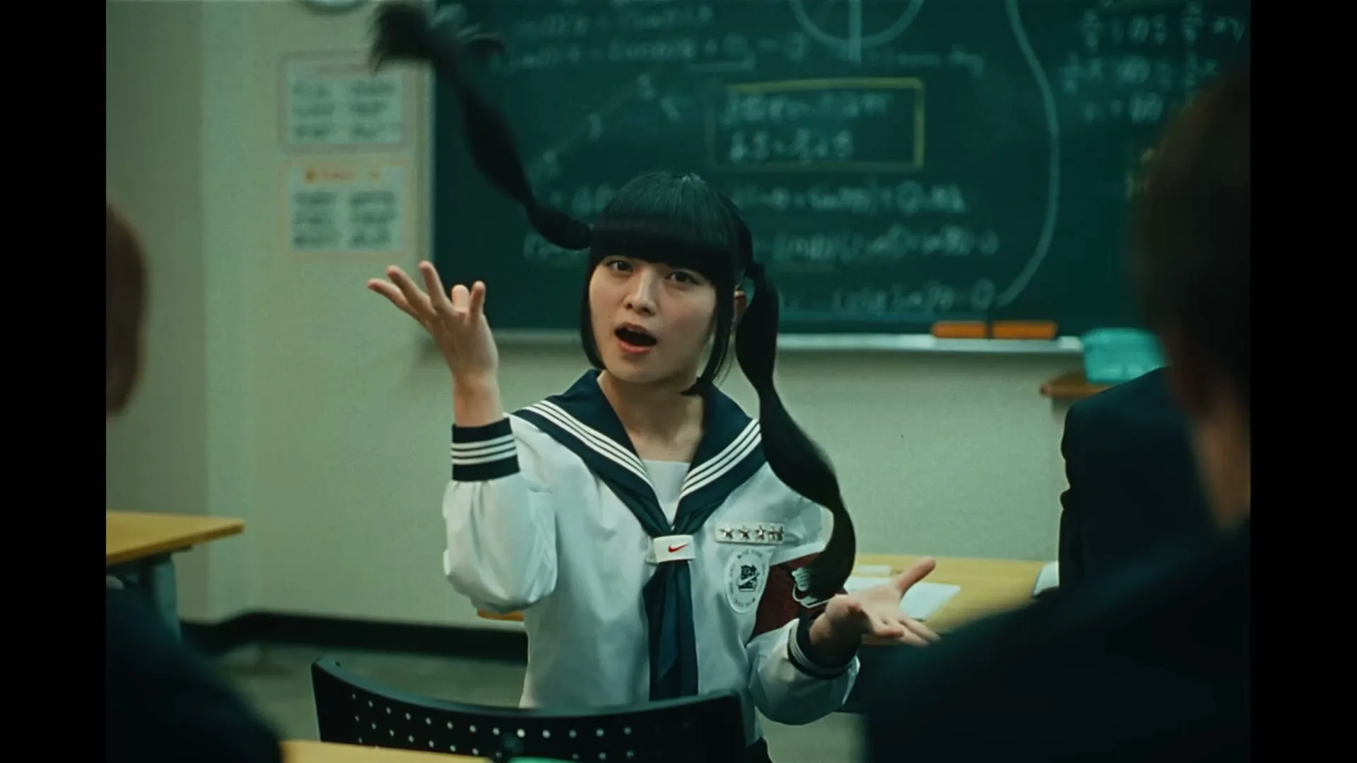A young Japanese woman with pigtails and a school uniform sits at a desk in a classroom, gesturing with her hands.