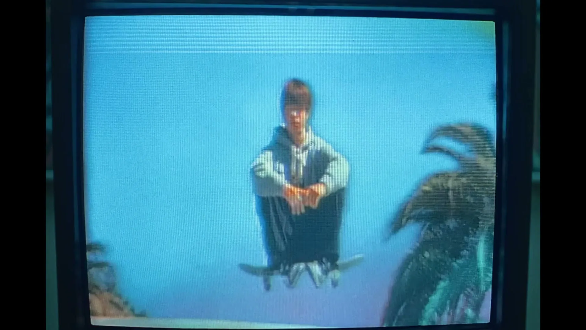 A young male skateboarder in a grey hoodie and dark pants performs an aerial trick, floating above a ramp against a bright blue sky with palm trees, displayed on an old TV screen.