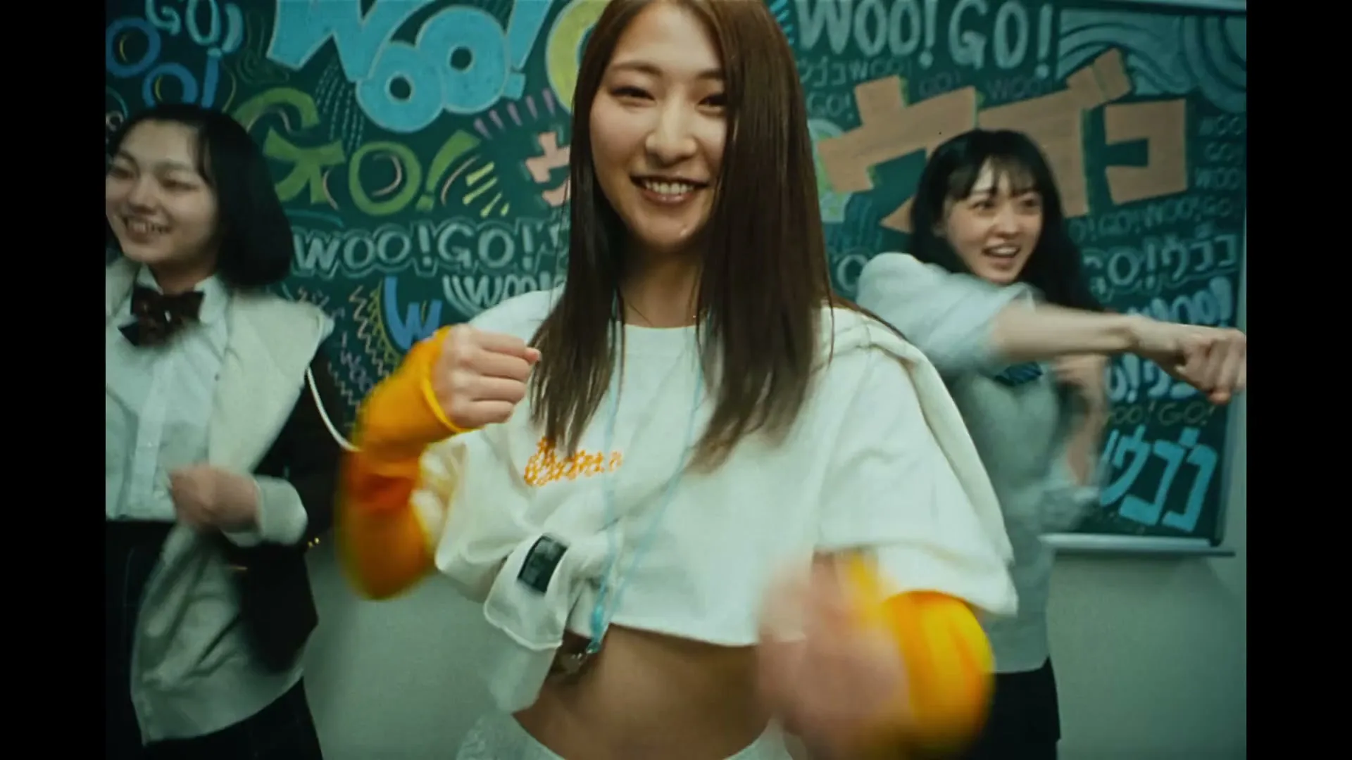 Three young Japanese women, one in a cropped white top and orange sleeves, dance energetically in front of a chalkboard with colorful graffiti.
