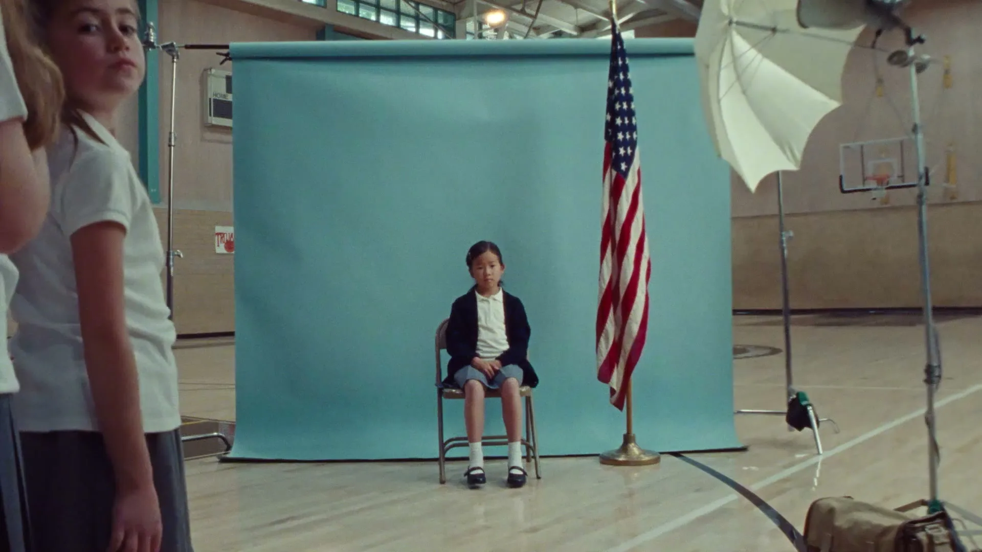 A young girl sits on a chair in front of a blue backdrop and an American flag, while another girl stands in the foreground in a school gym.