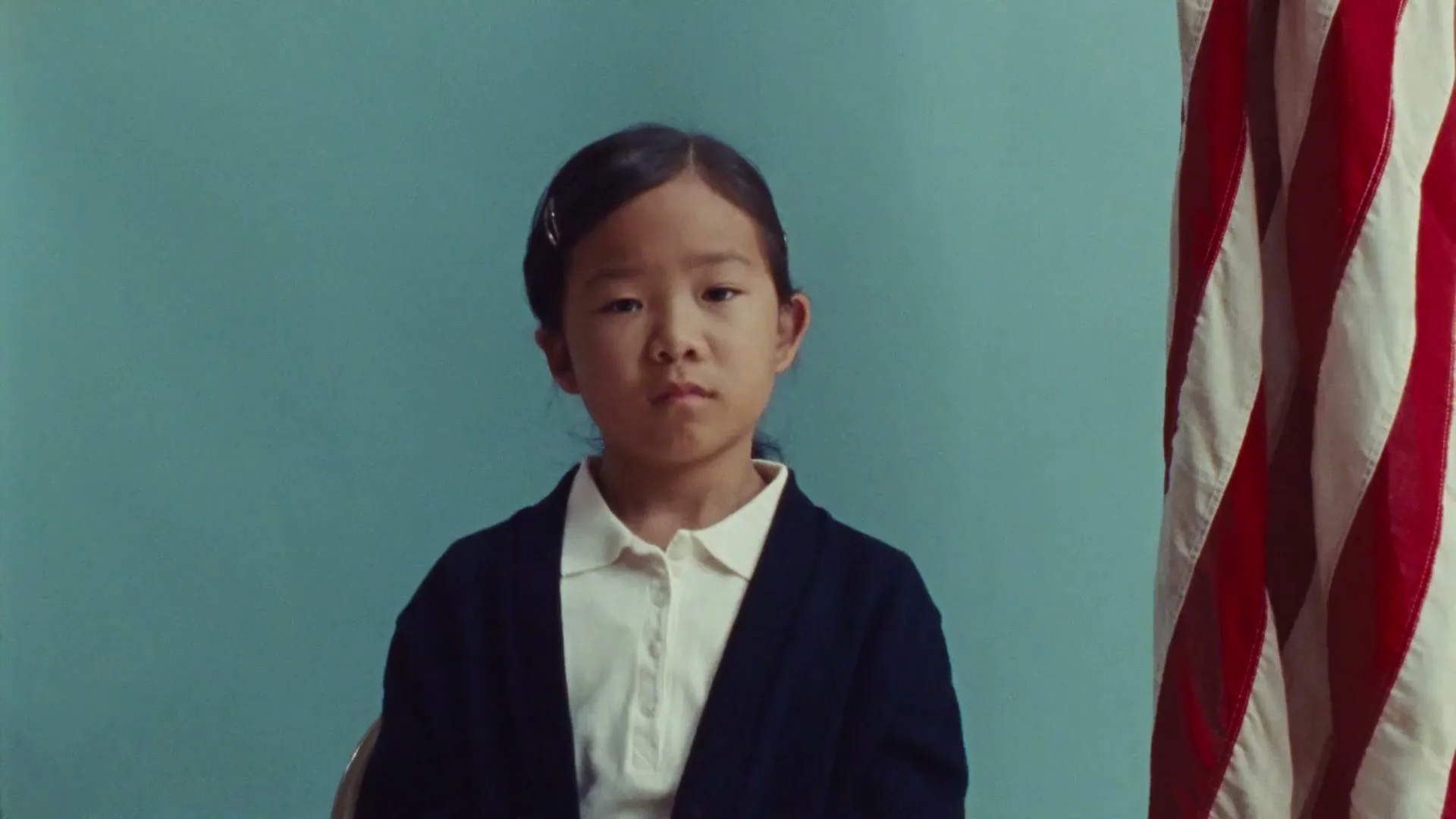 A young Asian girl with a serious expression sits in front of a blue wall and an American flag, then a hand reaches in front of her.