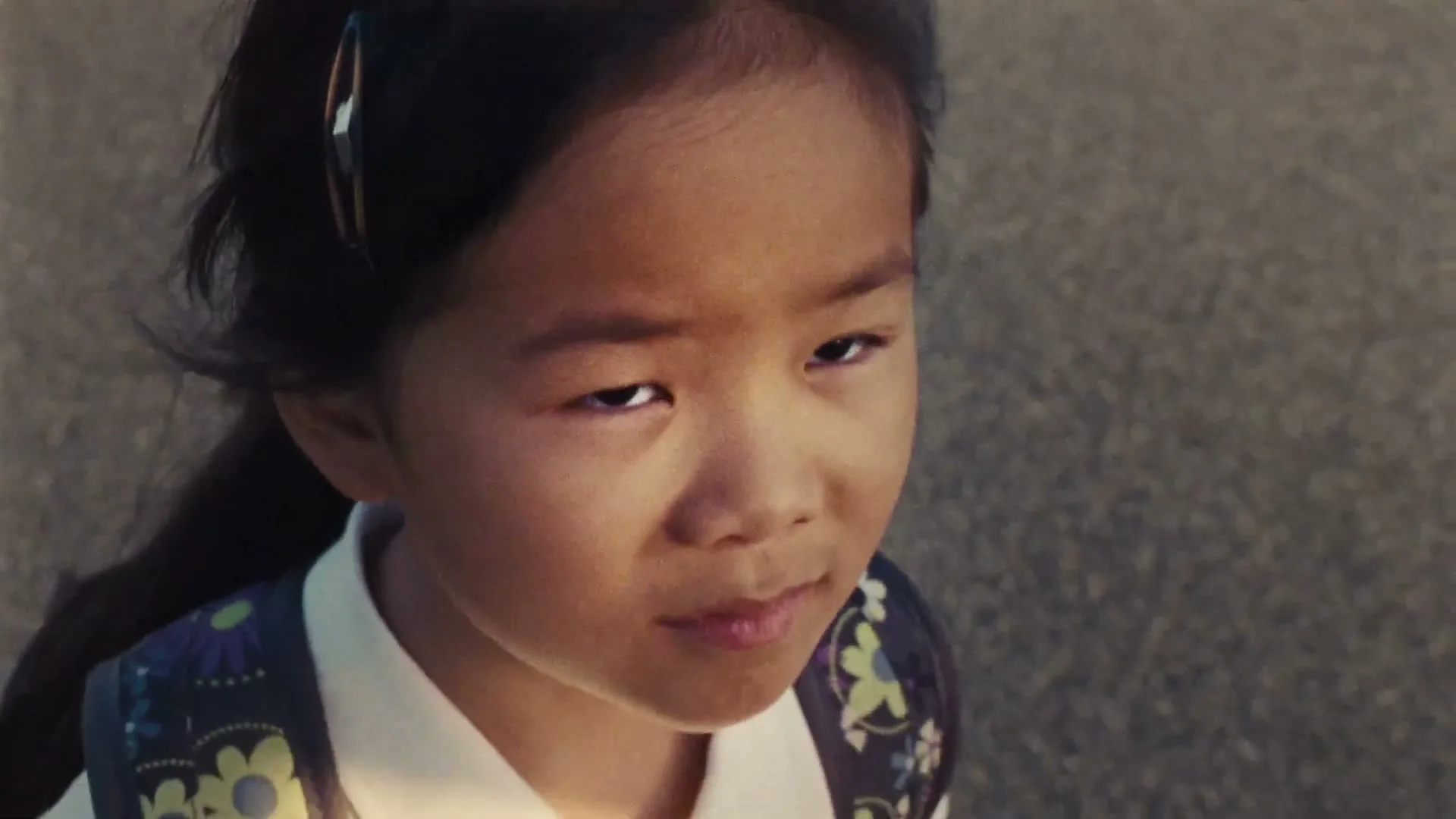 A young Asian girl with dark hair and a floral backpack looks up with a slight frown on her face, outdoors.