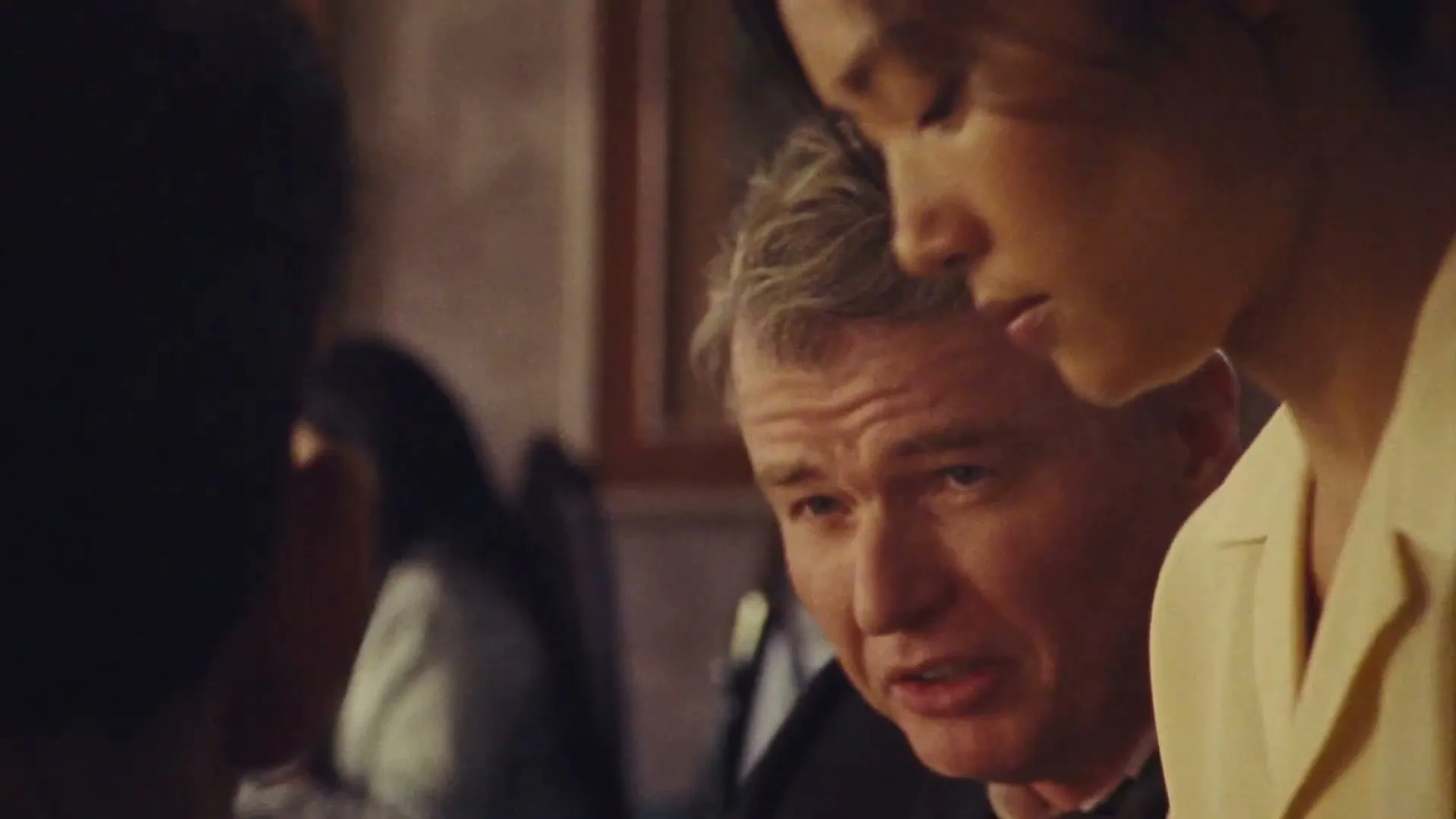 A young woman with dark hair in a ponytail leans over a man in a suit, who looks up at her with a concerned expression in an interior setting.