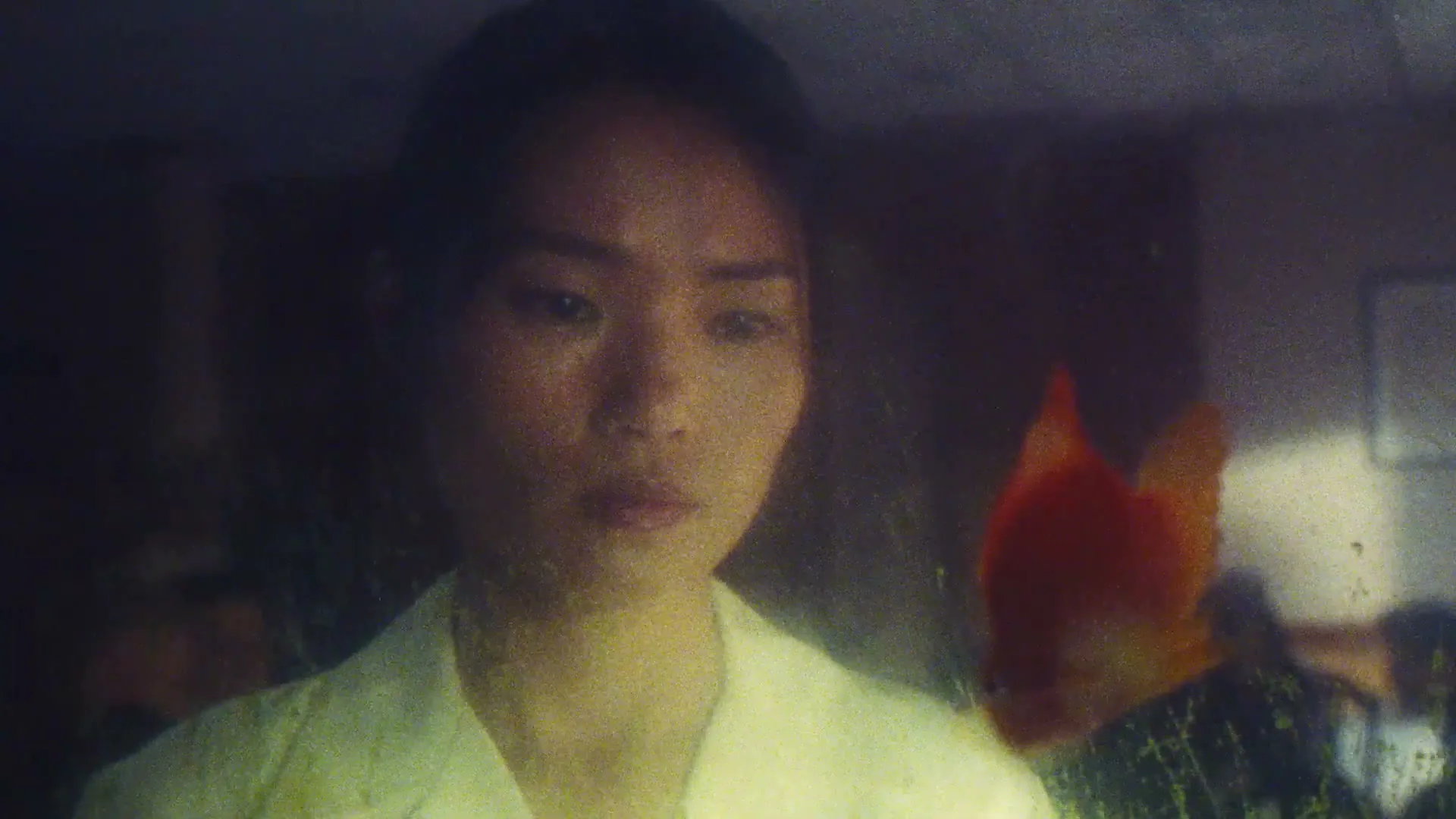 A young Asian woman in a light-colored shirt looks intently at a goldfish swimming in a tank, with a dark, blurry background.