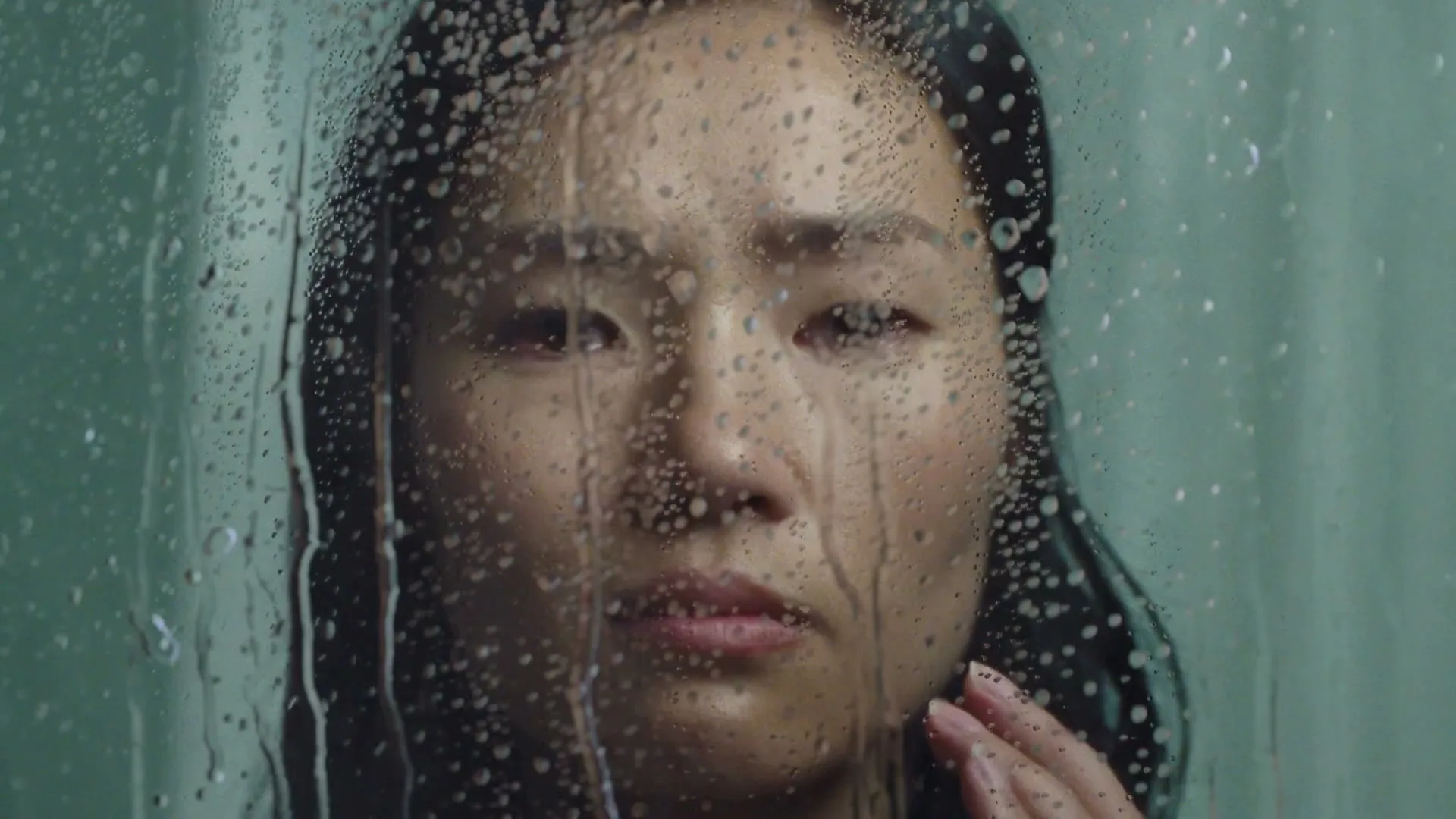 A woman with dark hair looks through a wet glass surface, her face appearing distressed as water droplets run down the glass.