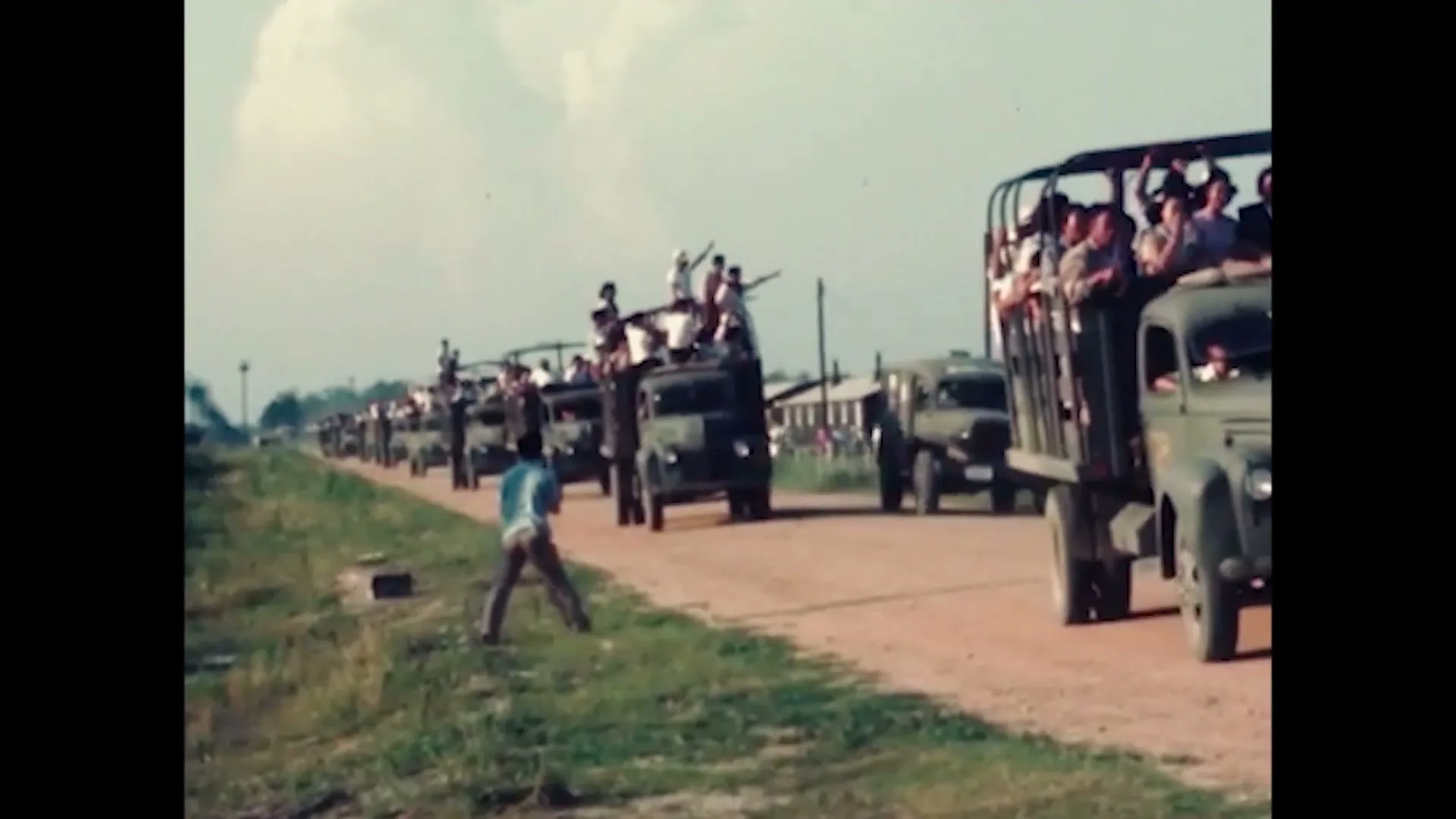 A long line of military trucks filled with people drives down a dirt road, while a man in a blue shirt stands by the roadside.