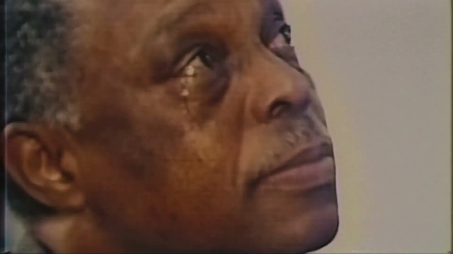A close-up shot of an older Black man with a mustache, looking up with tears streaming down his face, in an interior setting.