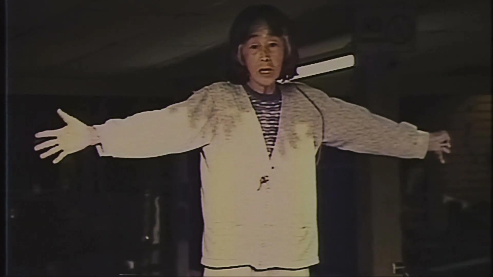 A person with long hair and a striped shirt stands with arms outstretched, looking distressed in a dark, grainy room.
