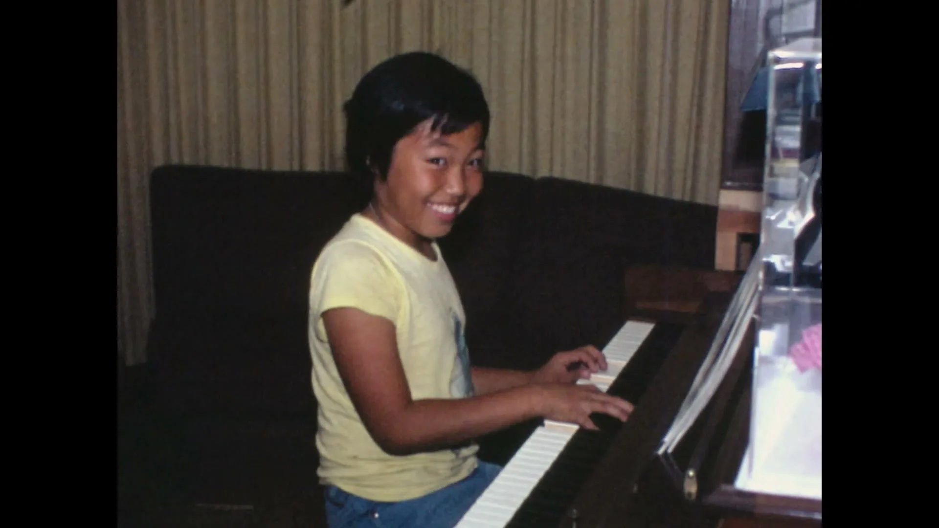 A young Asian child with short dark hair, wearing a yellow t-shirt and blue jeans, plays the piano in a living room, then looks up and smiles at the camera.
