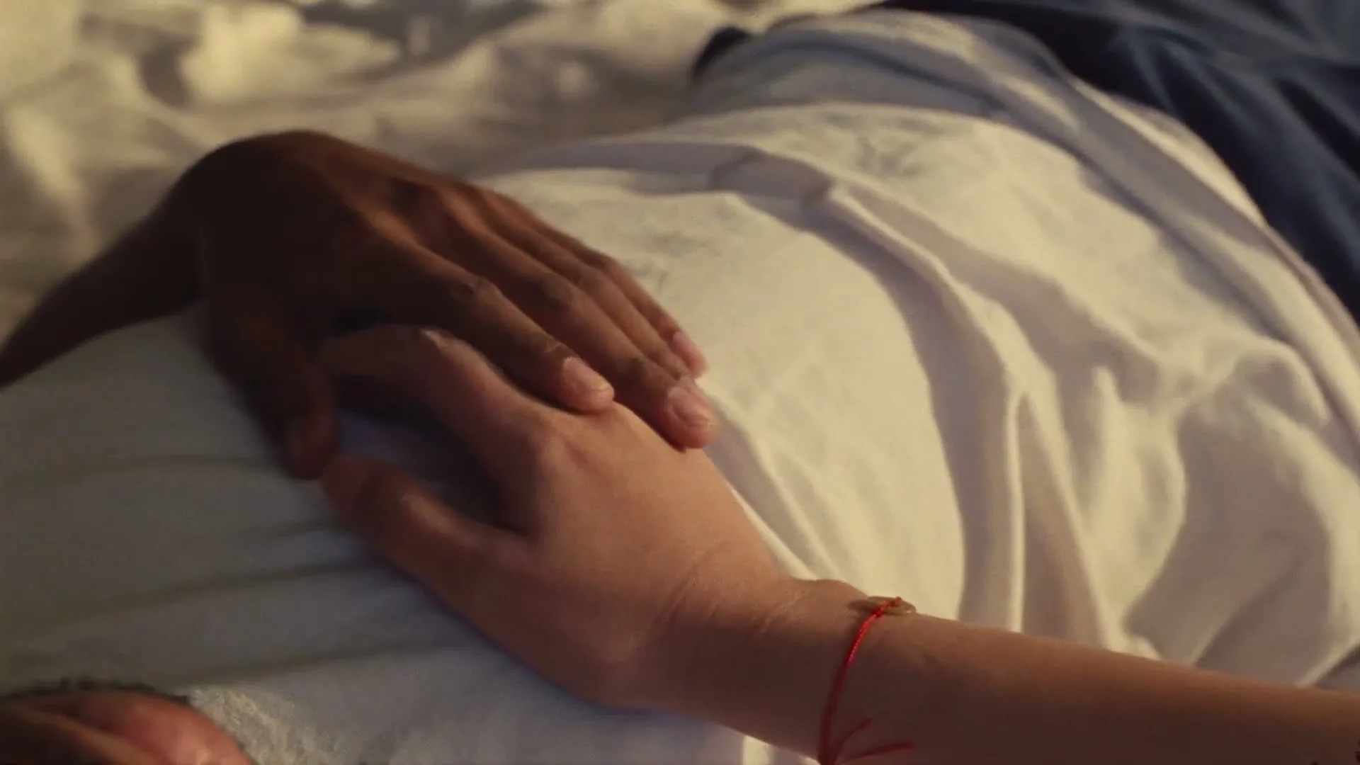 A close-up shot shows two hands, one with darker skin and one with lighter skin, gently touching on a white sheet.