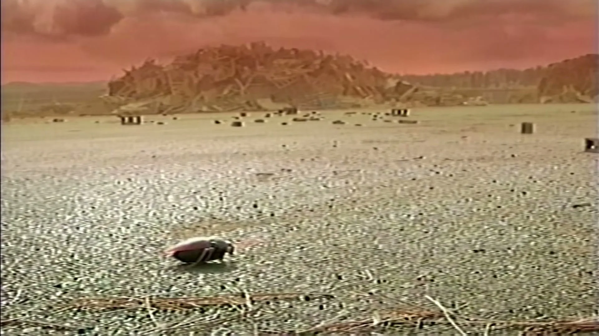 A wide shot of a desolate, rocky landscape under a red sky, with a small, dark object moving across the foreground.