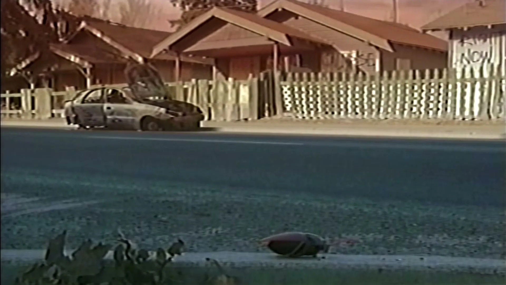 A dark, blurry object rolls across a sidewalk in front of a broken-down car and houses with picket fences, under a reddish sky.