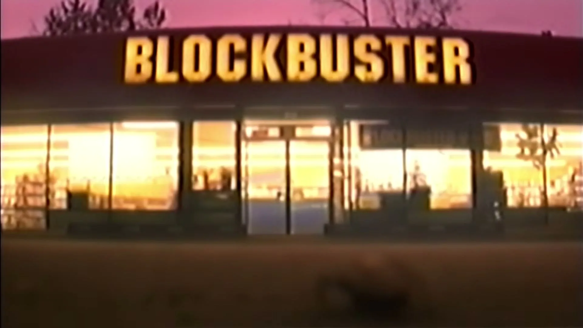 A cockroach crawls on the ground in front of a Blockbuster store at dusk, with the store's sign glowing brightly.