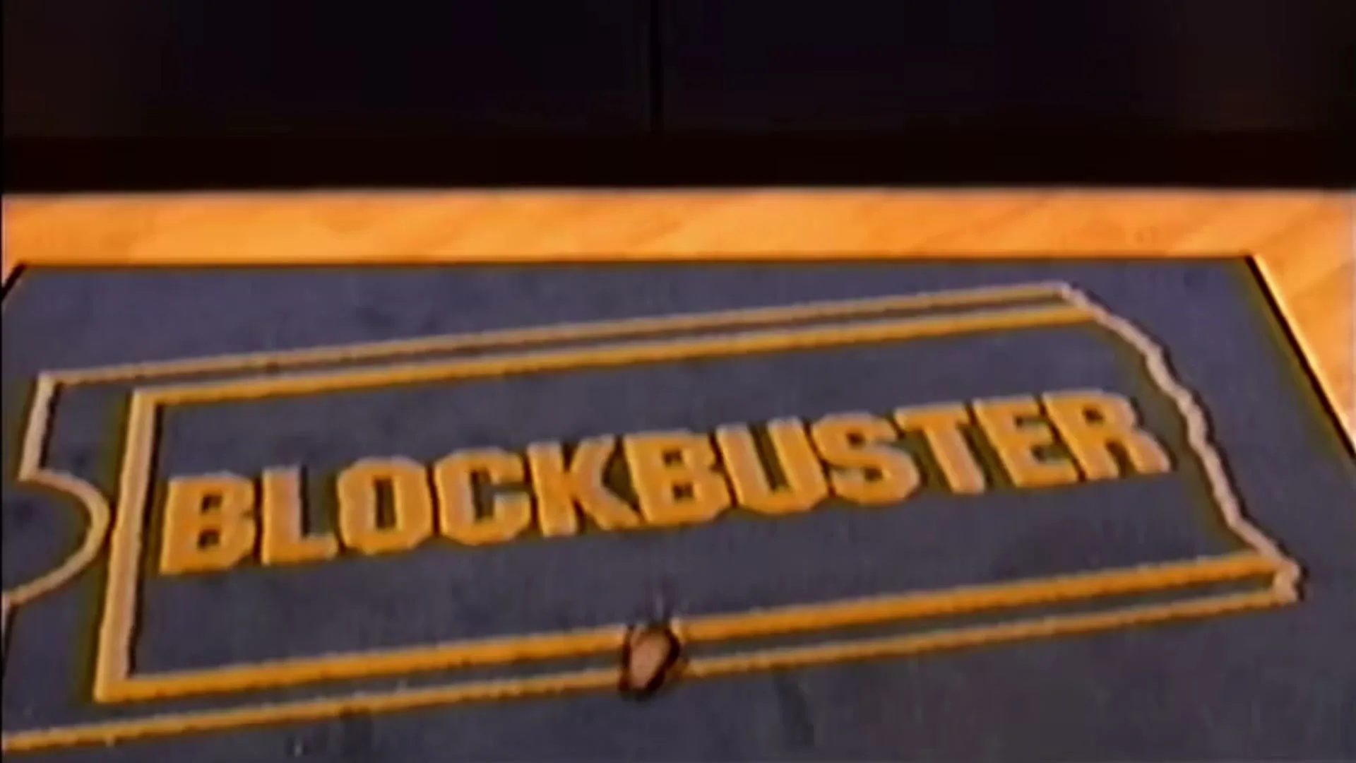 A camera pans down to reveal a blue Blockbuster logo mat on a light wooden floor, with a dark doorway in the foreground.