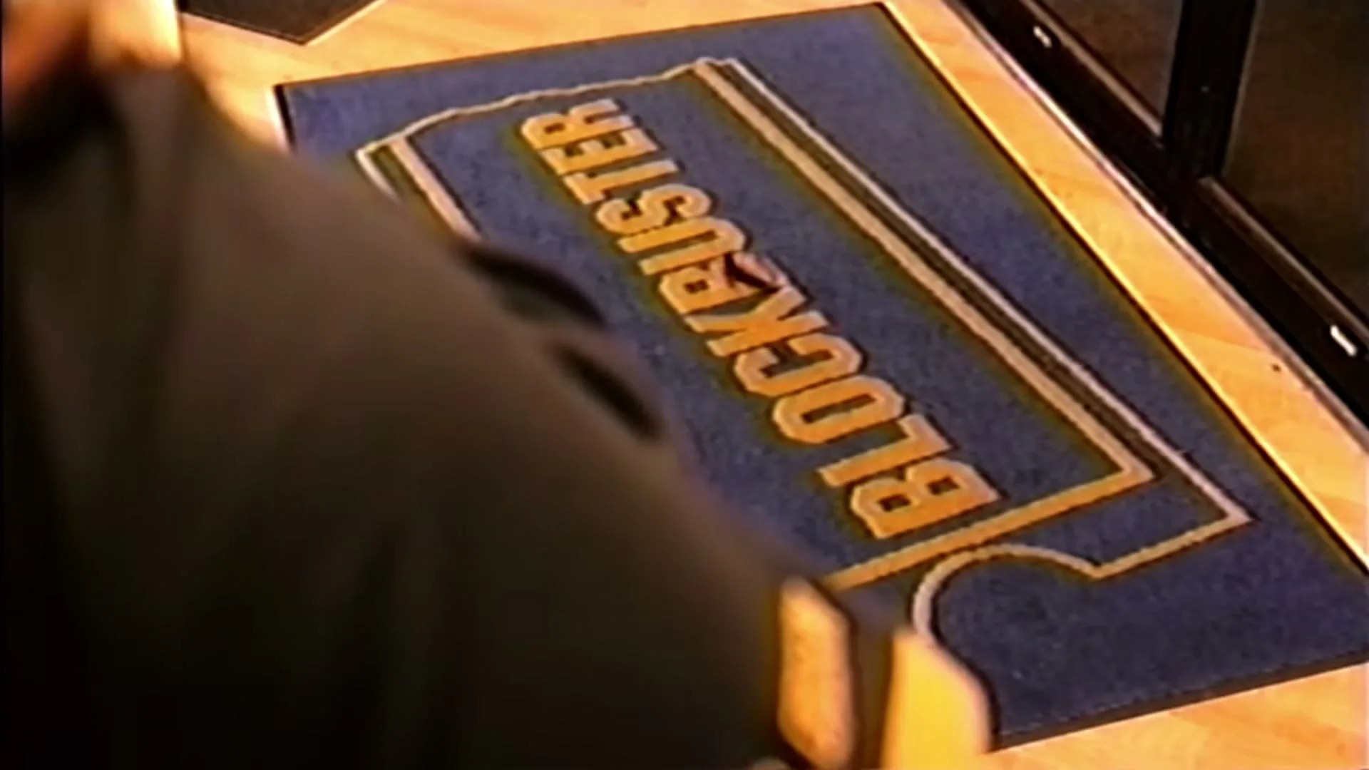 A person's arm and shoulder are visible as they step on a blue Blockbuster logo doormat inside a store.