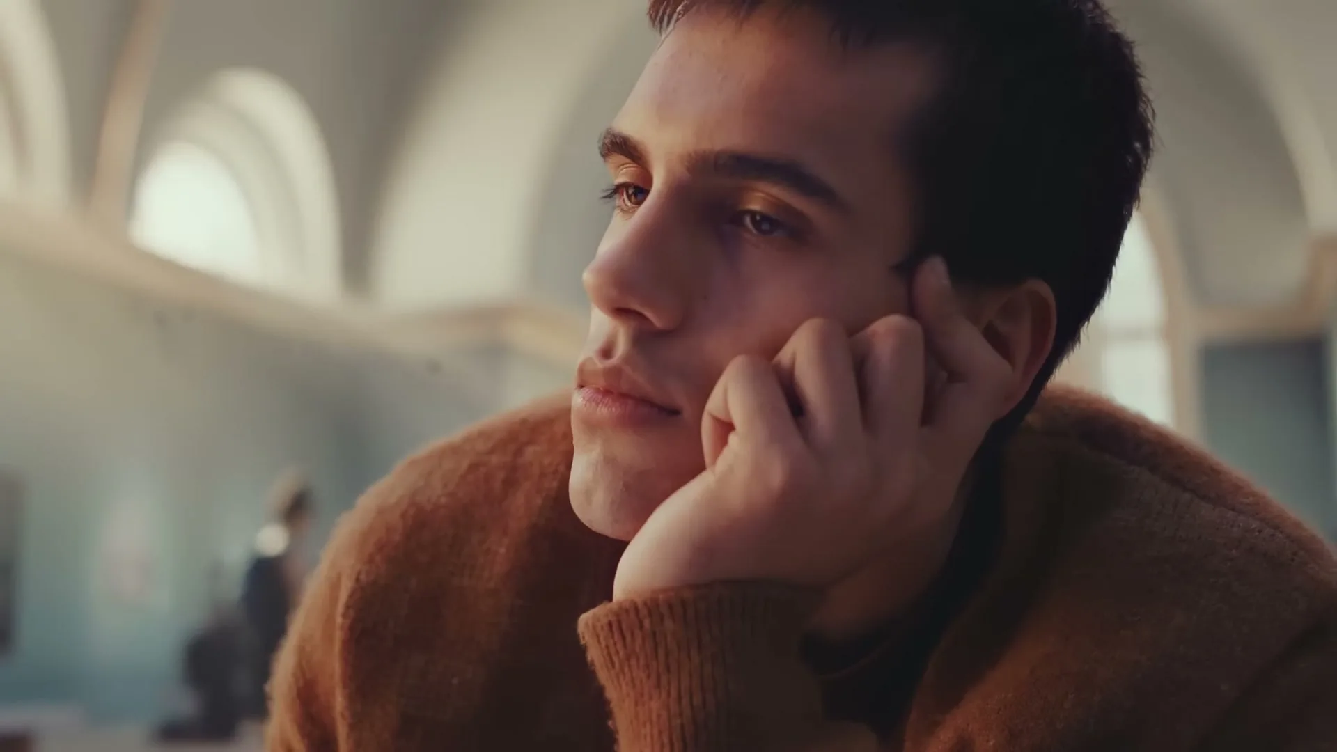 A young man with dark hair and a brown sweater rests his chin on his hand, looking thoughtfully to the side in a bright, airy museum.