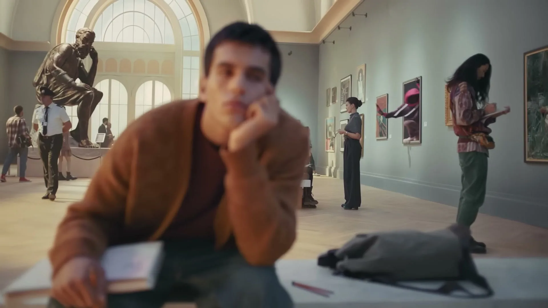 A young man in a brown cardigan sits bored in a museum, resting his head on his hand, as other patrons look at art.