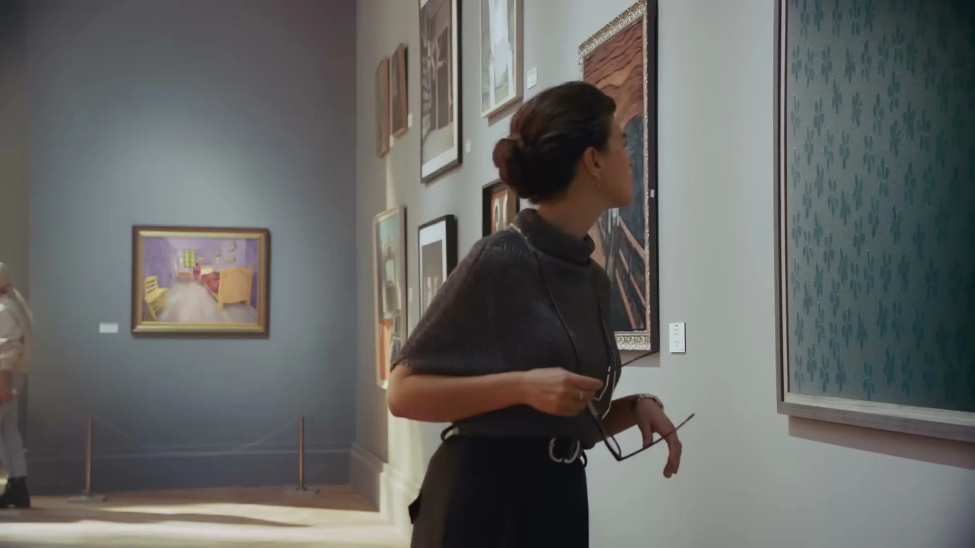 A woman in a dark top and skirt holds glasses while looking intently at a painting in an art gallery.