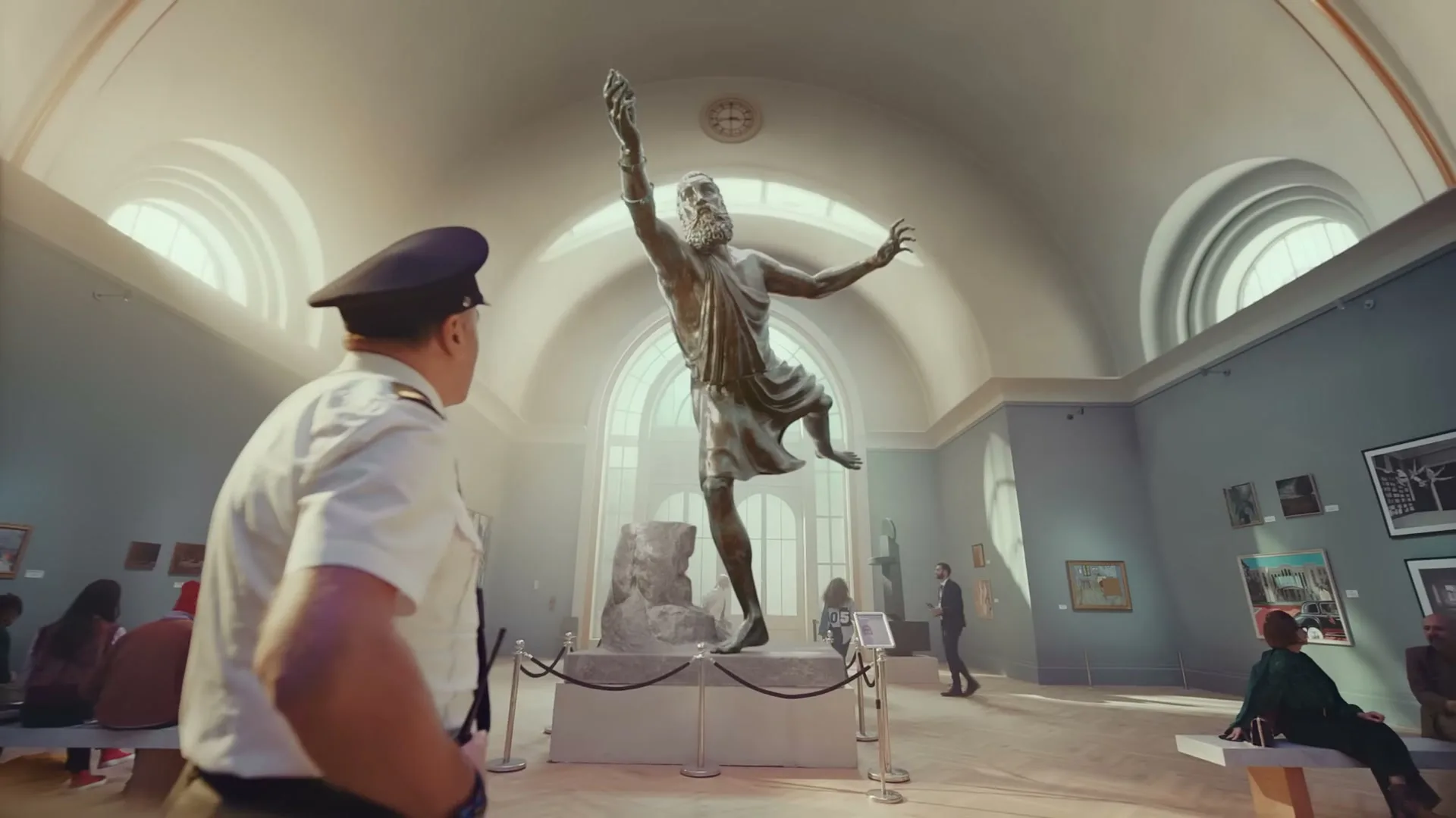 A museum guard in a white uniform and hat walks past a large bronze statue of a man with his arms raised in a brightly lit museum gallery.