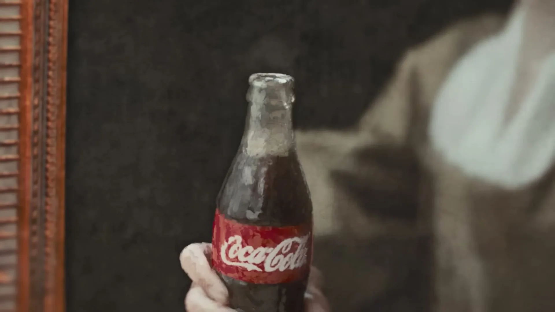 A painted hand holds a Coca-Cola bottle as another painted hand uses a bottle opener to remove the cap, releasing a puff of smoke.