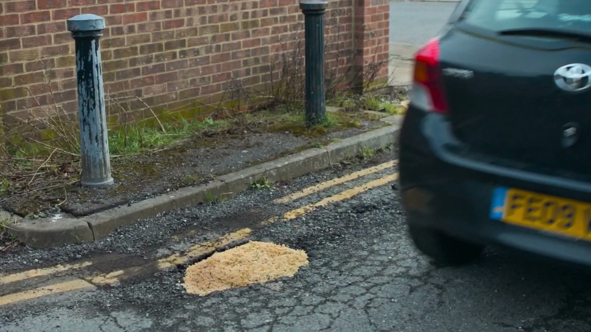 A black car drives past a pothole filled with noodles on a cracked asphalt road next to a brick wall.