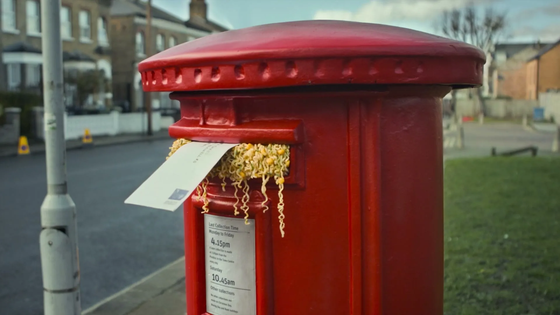 A red British post box on a suburban street has instant noodles spilling out of its letter slot, with a letter stuck in the noodles.