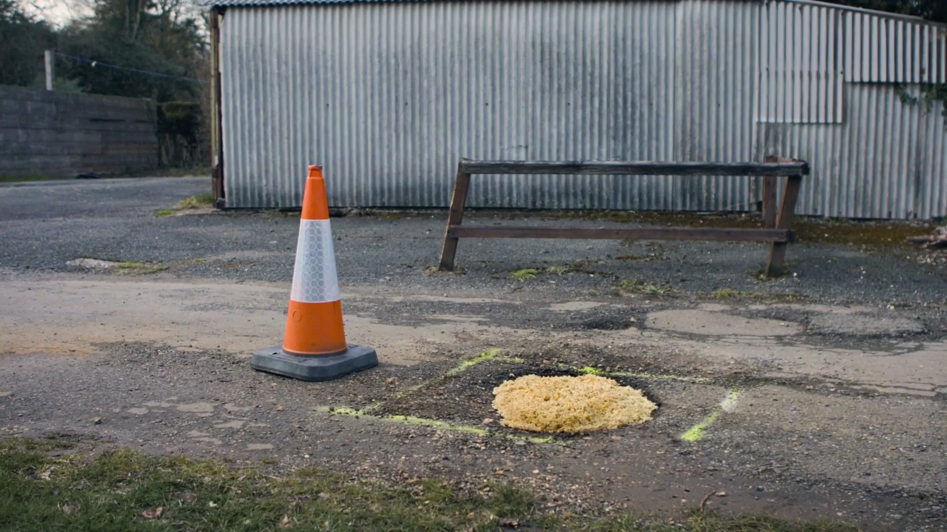 A traffic cone stands next to a pothole filled with Pot Noodle on a gravel road in front of a corrugated metal building.
