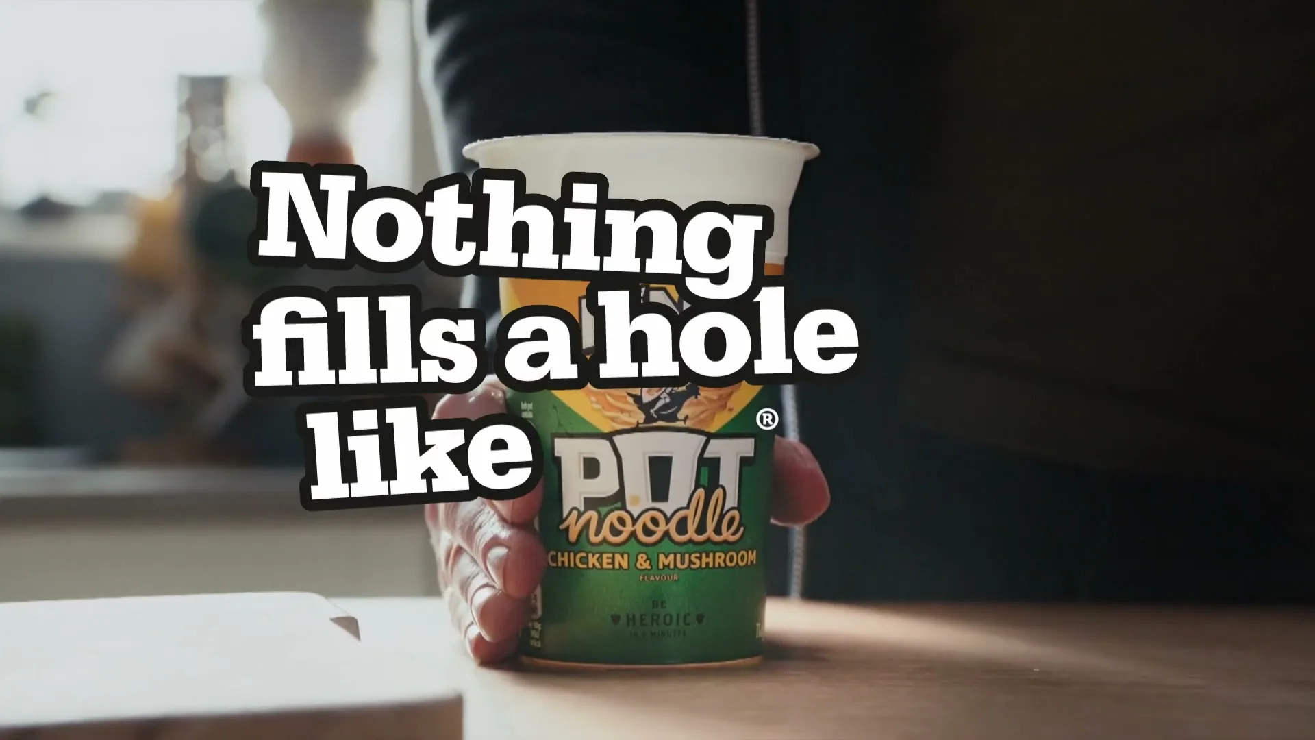 A person's hand places a Pot Noodle cup on a wooden table in a kitchen, with the tagline "Nothing fills a hole like Pot Noodle" superimposed.