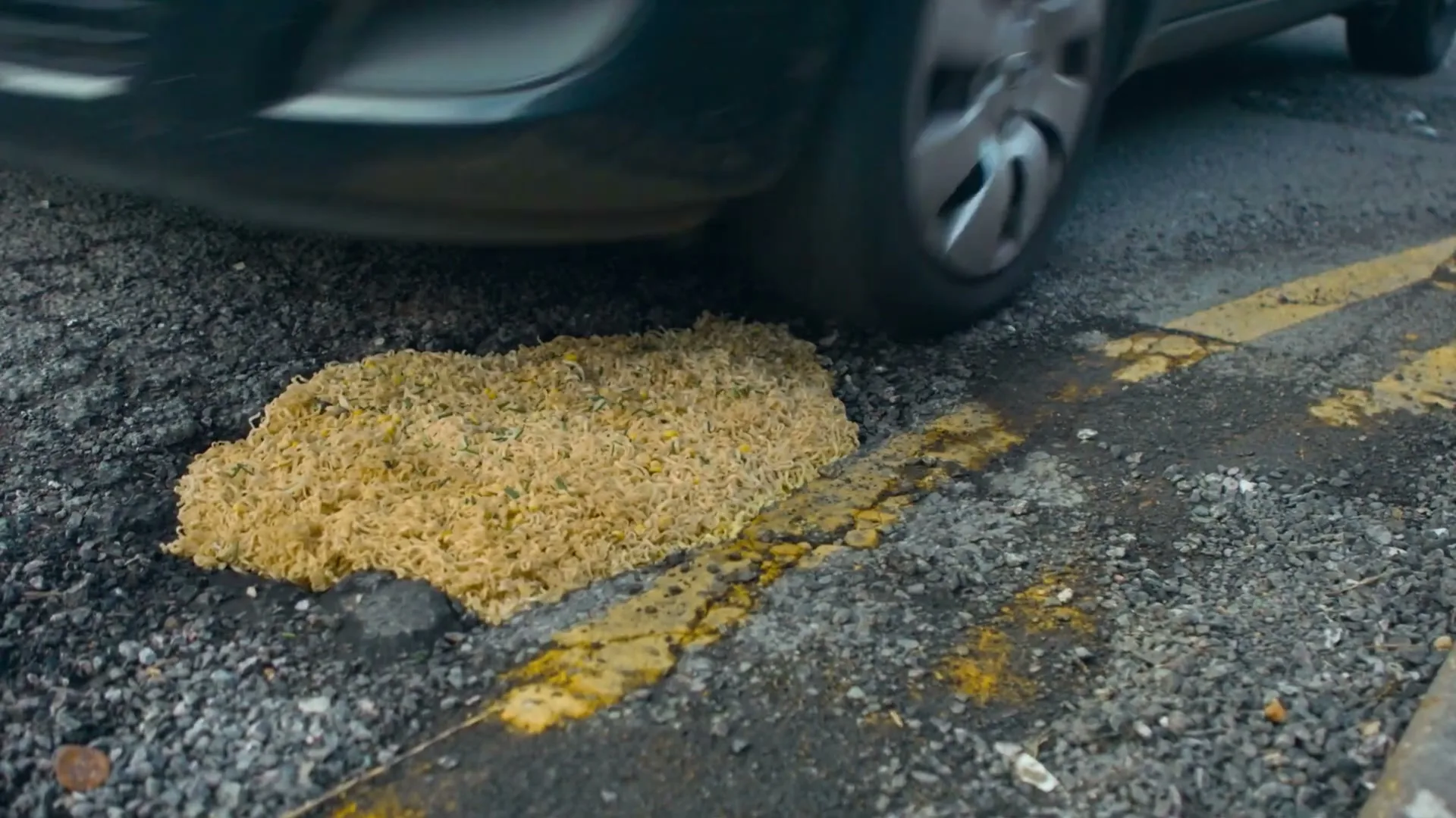 A pothole filled with dry instant noodles on an asphalt road with a car tire driving over it, splashing the noodles.