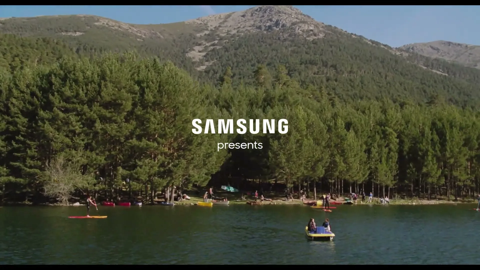 An aerial wide shot shows a beautiful lake surrounded by a lush green forest and mountains, with people enjoying various water activities on a sunny day.