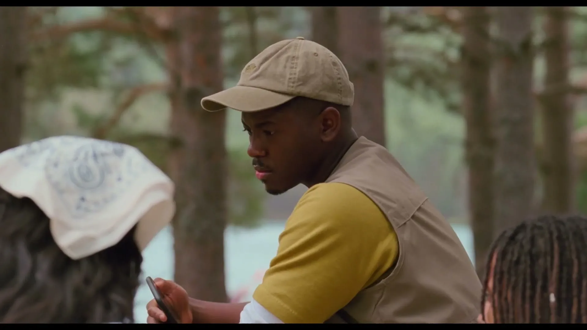 A young man in a cap and vest looks down at his phone while sitting outdoors with friends in a forest setting.