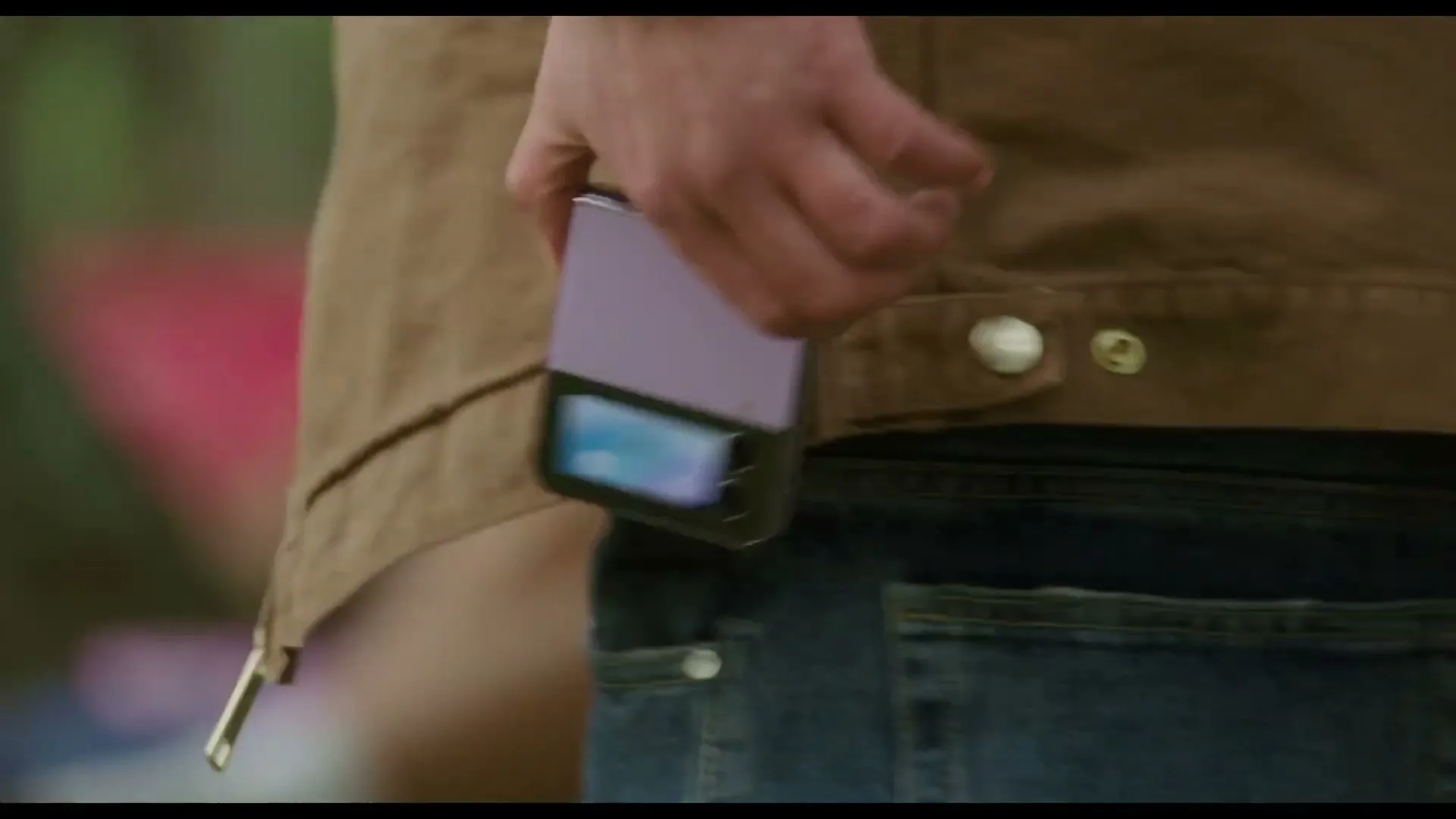 A person in a brown jacket and blue jeans pulls a purple Samsung Galaxy Z Flip phone from their back pocket, then holds it in their hands.