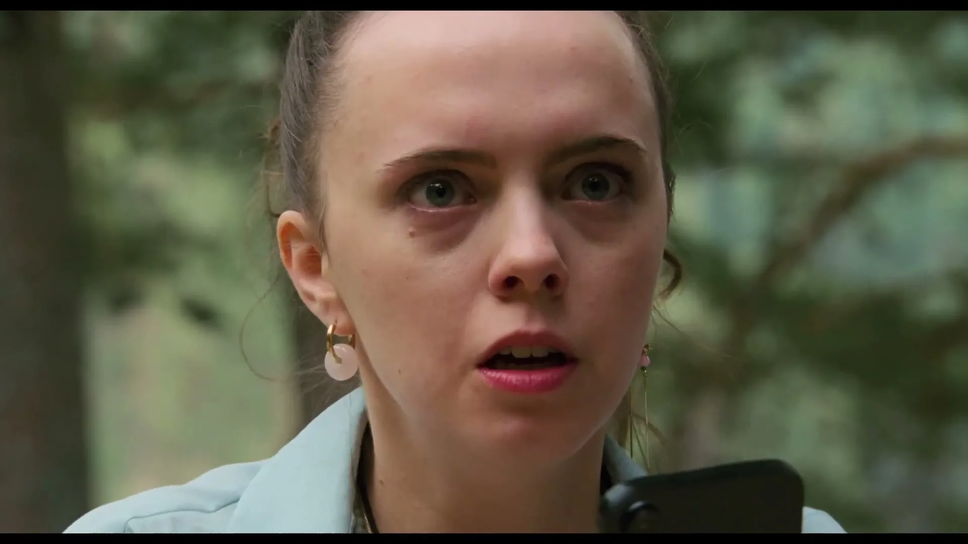 A young woman with light brown hair tied back, wearing a light blue jacket and earrings, looks shocked while holding a phone in a forest.