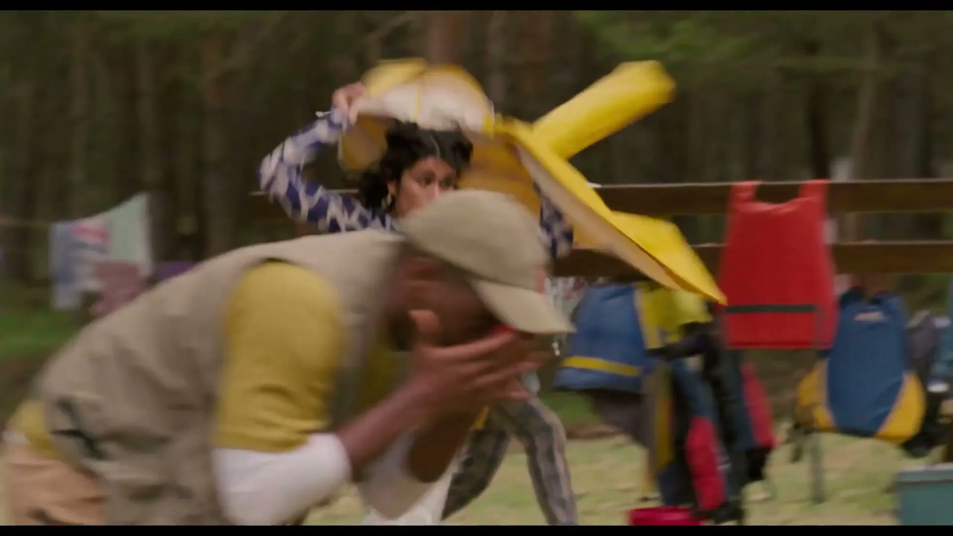 A group of young adults at a summer camp run away from something unseen, covering their faces and ducking, in a forest clearing.