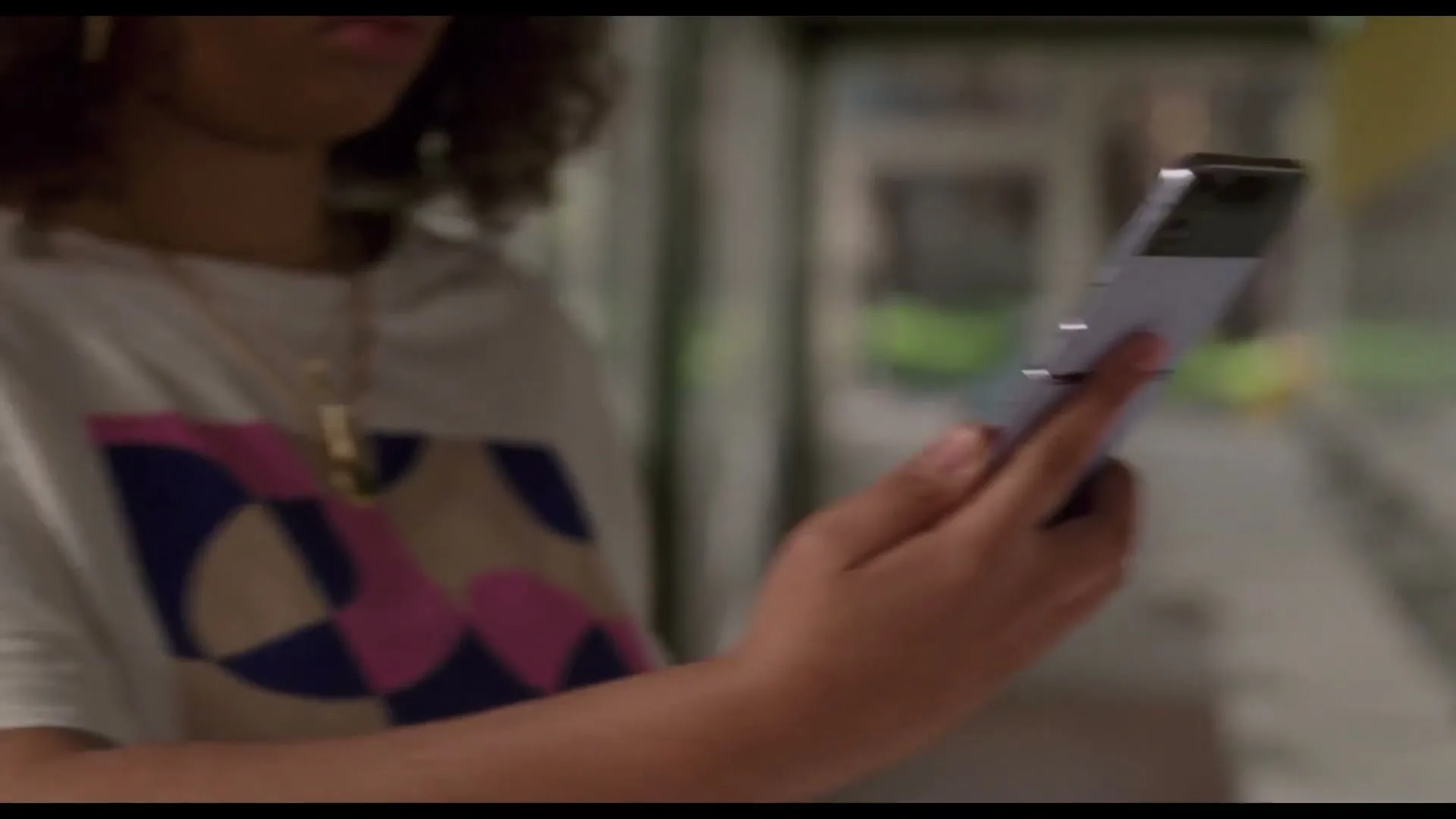 A young woman in a white t-shirt with a colorful graphic holds a purple flip phone, looking out a window at a man in a green vest running on a sunny lawn.