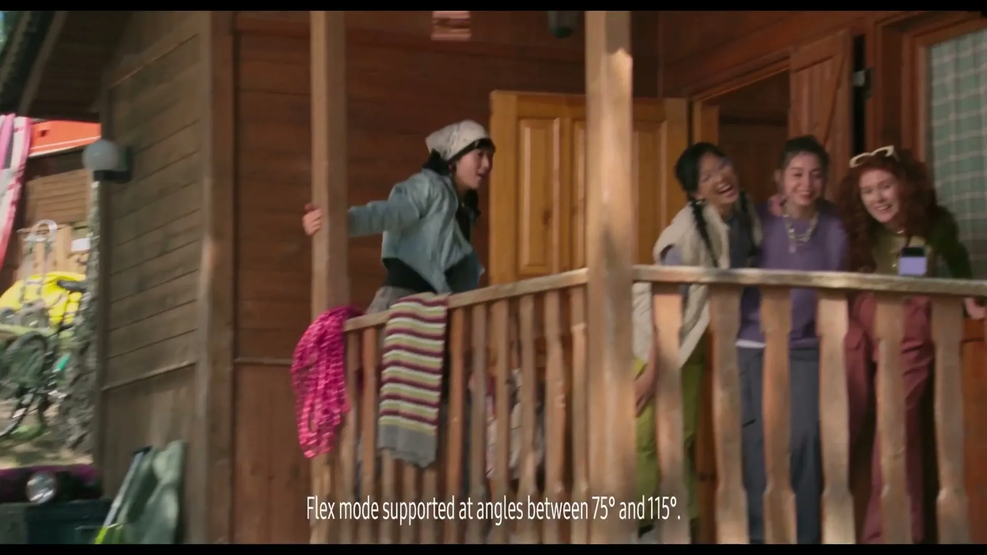 Four young women on a wooden porch, one jumps over the railing while the others laugh, at a rustic cabin.