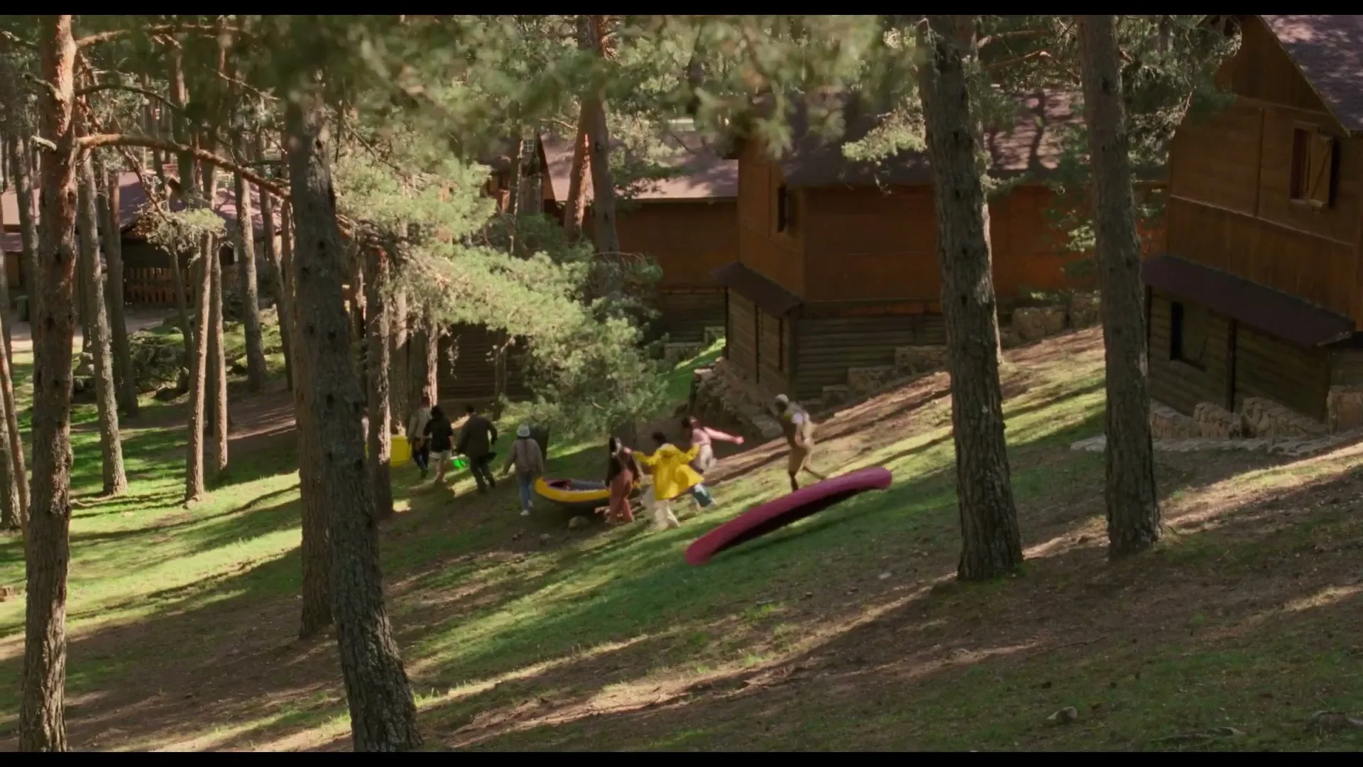 A group of people in a forest run away from a red canoe and a yellow inflatable boat that are sliding down a grassy hill towards wooden cabins.