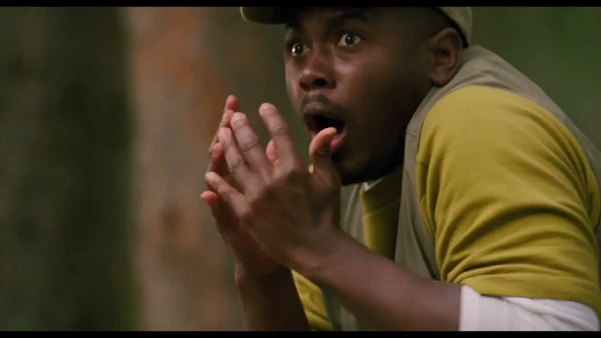 A man in a cap and vest looks shocked, holding his hands to his mouth, in a forest with trees and another person in the background.